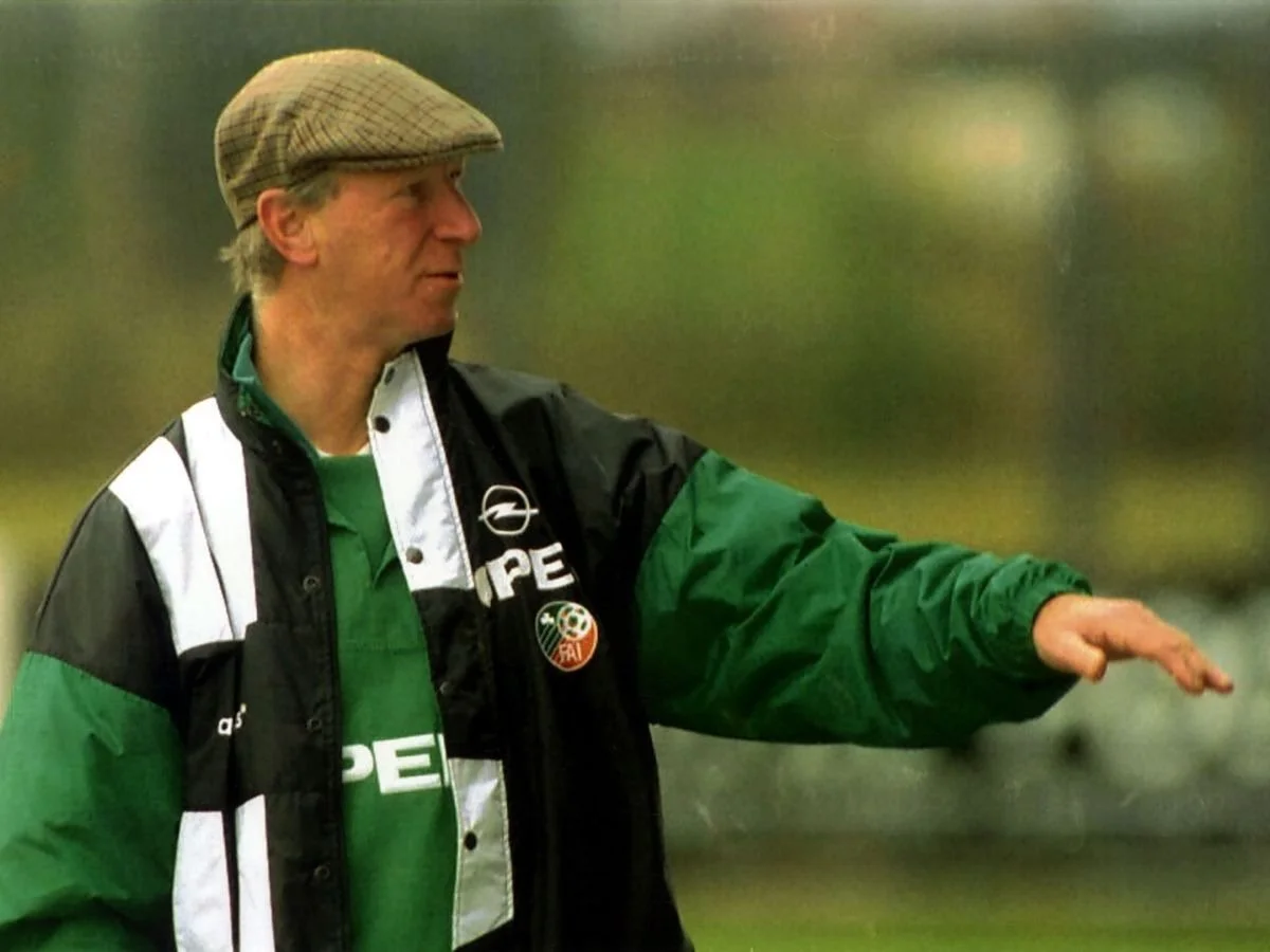 A man wearing a brown checkered cap, green shirt, and a black and green jacket with Opel and other logos, standing outdoors and gesturing with his right hand.