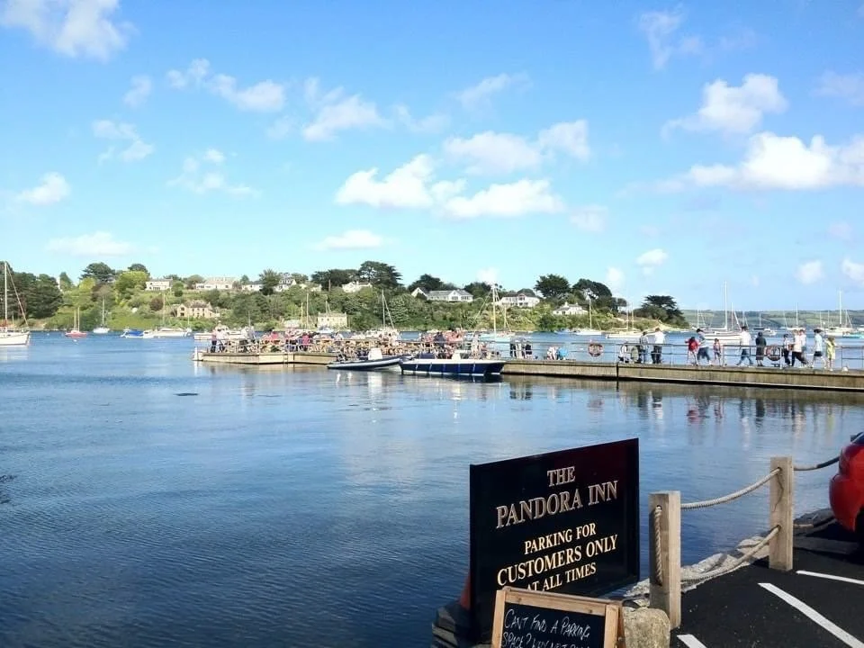 Harbor with boats docked, people walking along the pier, and a sign for The Pandora Inn, a parking lot for customers, and a scenic view of water and houses in the background.