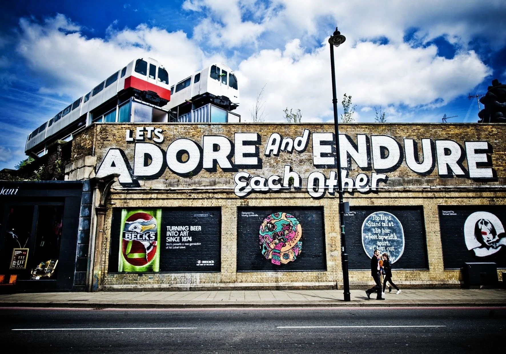 Street view of a brick building with large graffiti-style mural that says "Let's Adore and Endure Each Other" on the wall. There are two modern white and red train cars elevated above the building, a lamp post, and three people walking on the sidewal