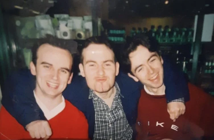 Three young men smiling and posing together indoors, two wearing red shirts and one in a checkered shirt with a blazer.