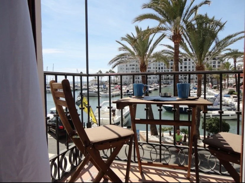 Balcony overlooking a marina with boats, palm trees, and a white building in the background, with a small wooden table and two chairs.