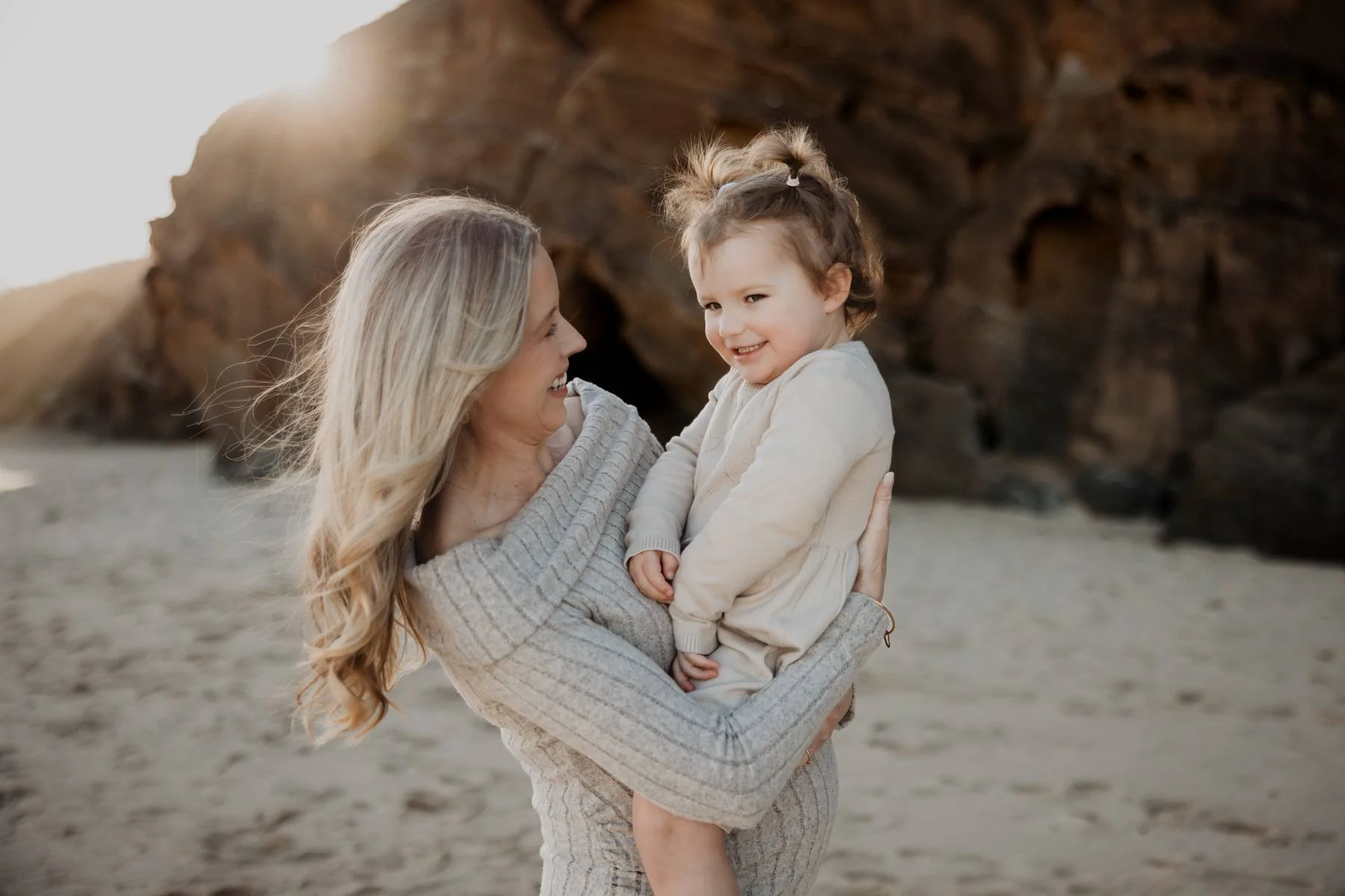 A woman holding a young girl at the beach with rock formations and the sun in the background.