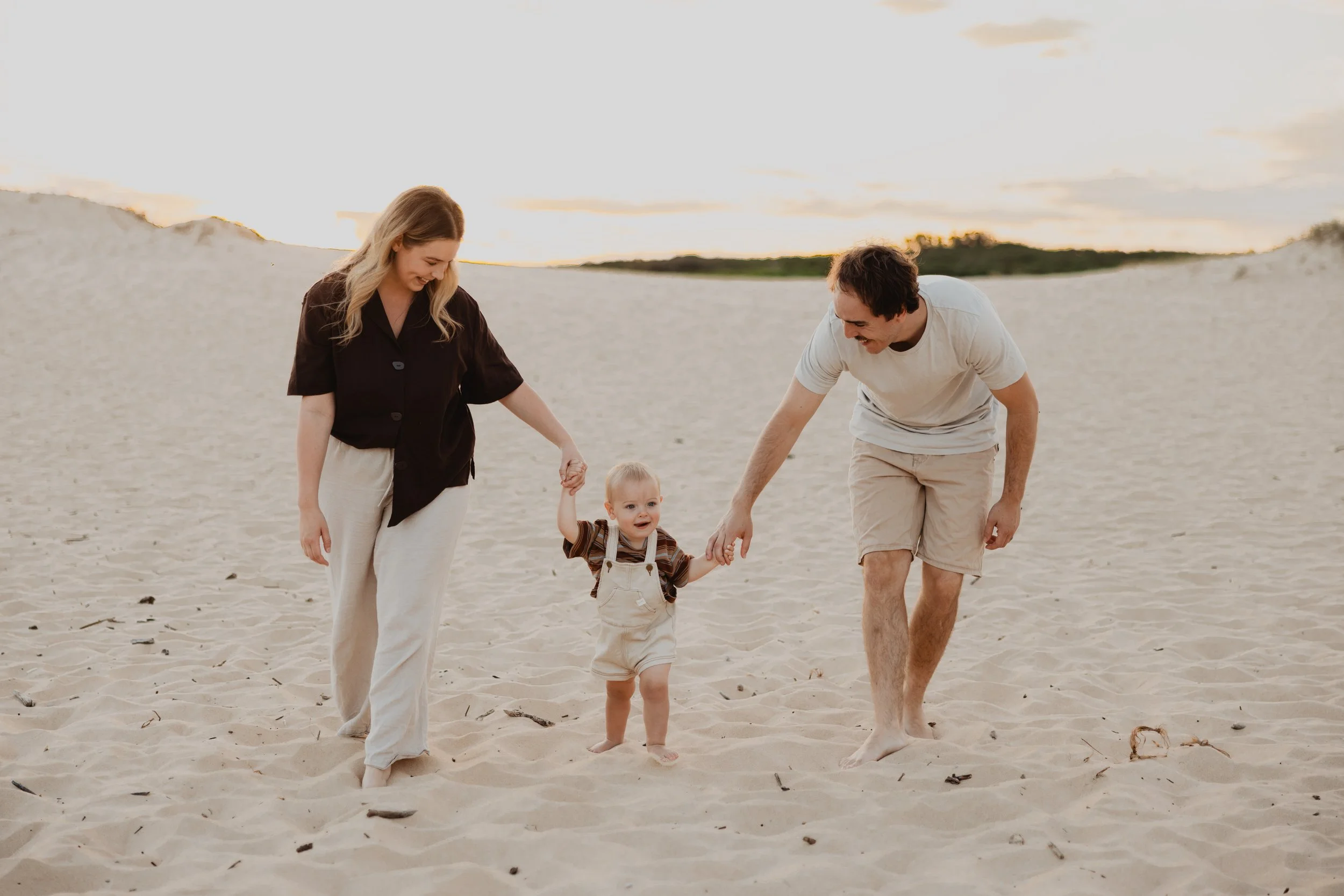 A family of three on a sandy beach at sunset, with a woman, a man, and a young child holding hands and smiling.