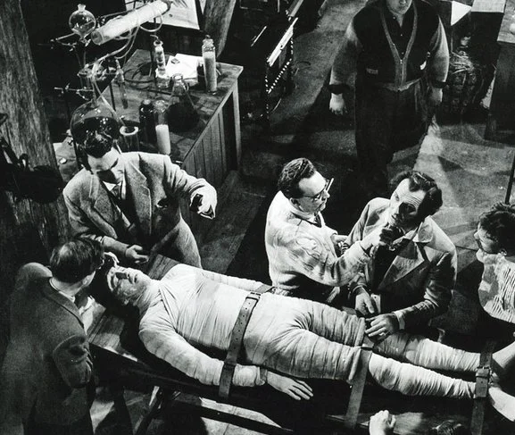 On the set of The Curse of Frankenstein, Cushing has his makeup touched up while Lee's creature lies strapped to the table.