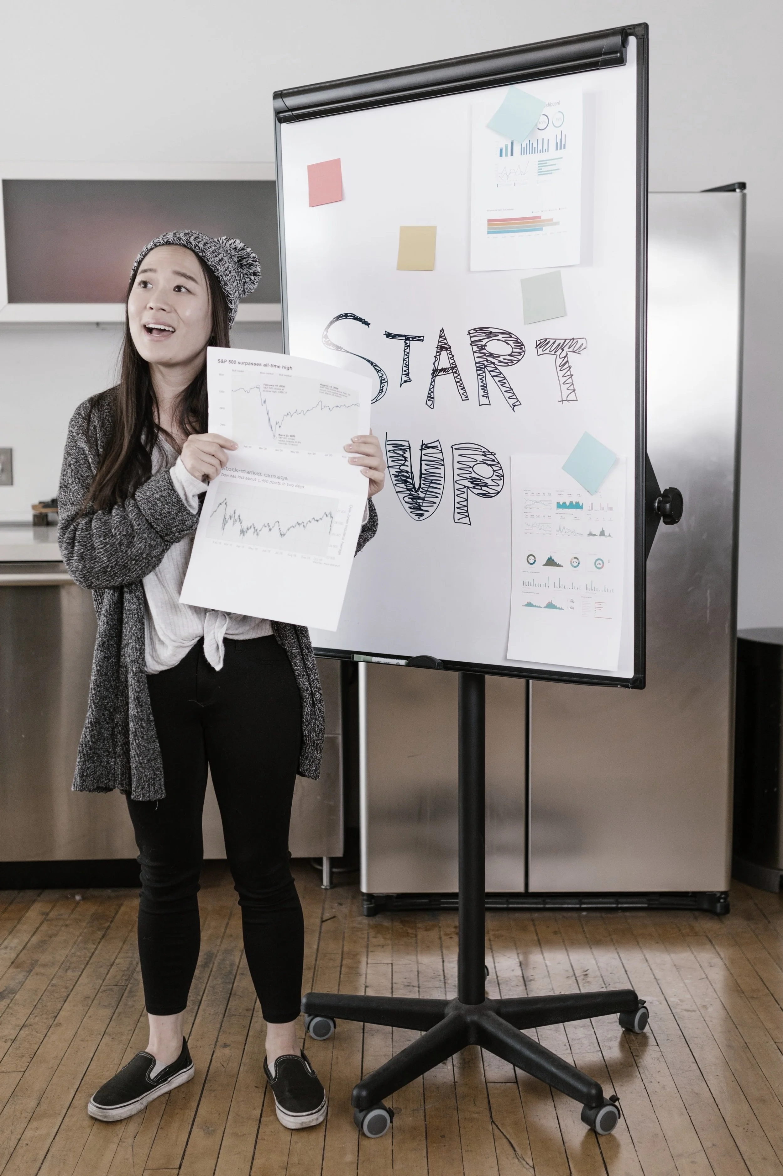 Woman giving a presentation in front of a whiteboard with "START UP" written on it, holding graphs and charts. She is wearing a beanie and casual outfit.