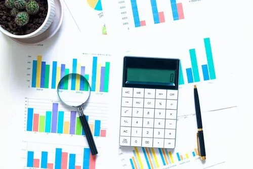 Business charts and graphs on papers, a calculator, pen, a magnifying glass, and a potted plant on a desk.