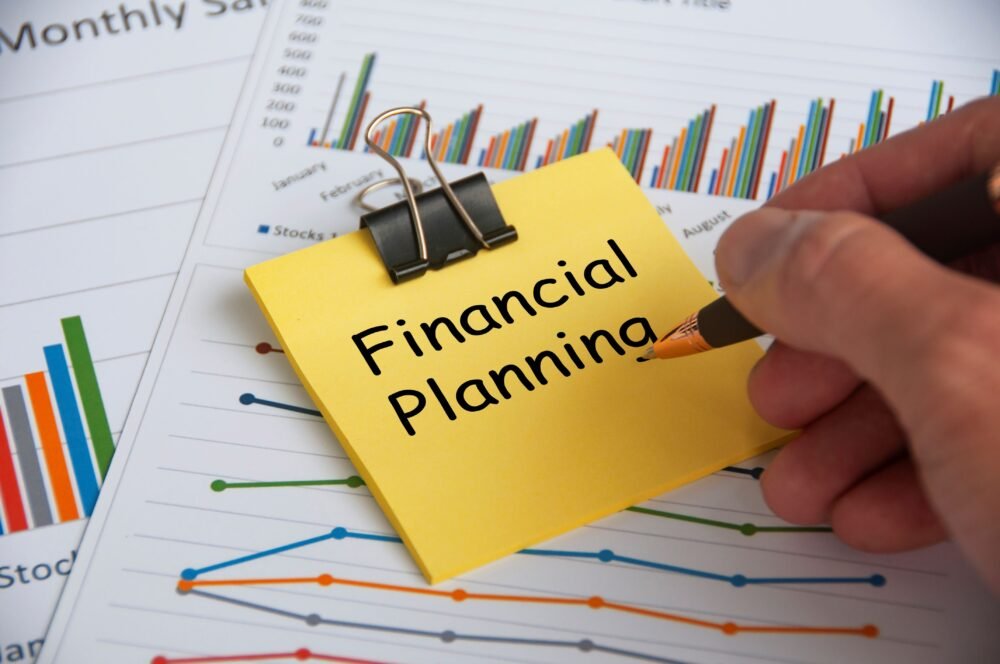 A person's hand writing on a yellow sticky note labeled 'Financial Planning' with charts and graphs in the background.