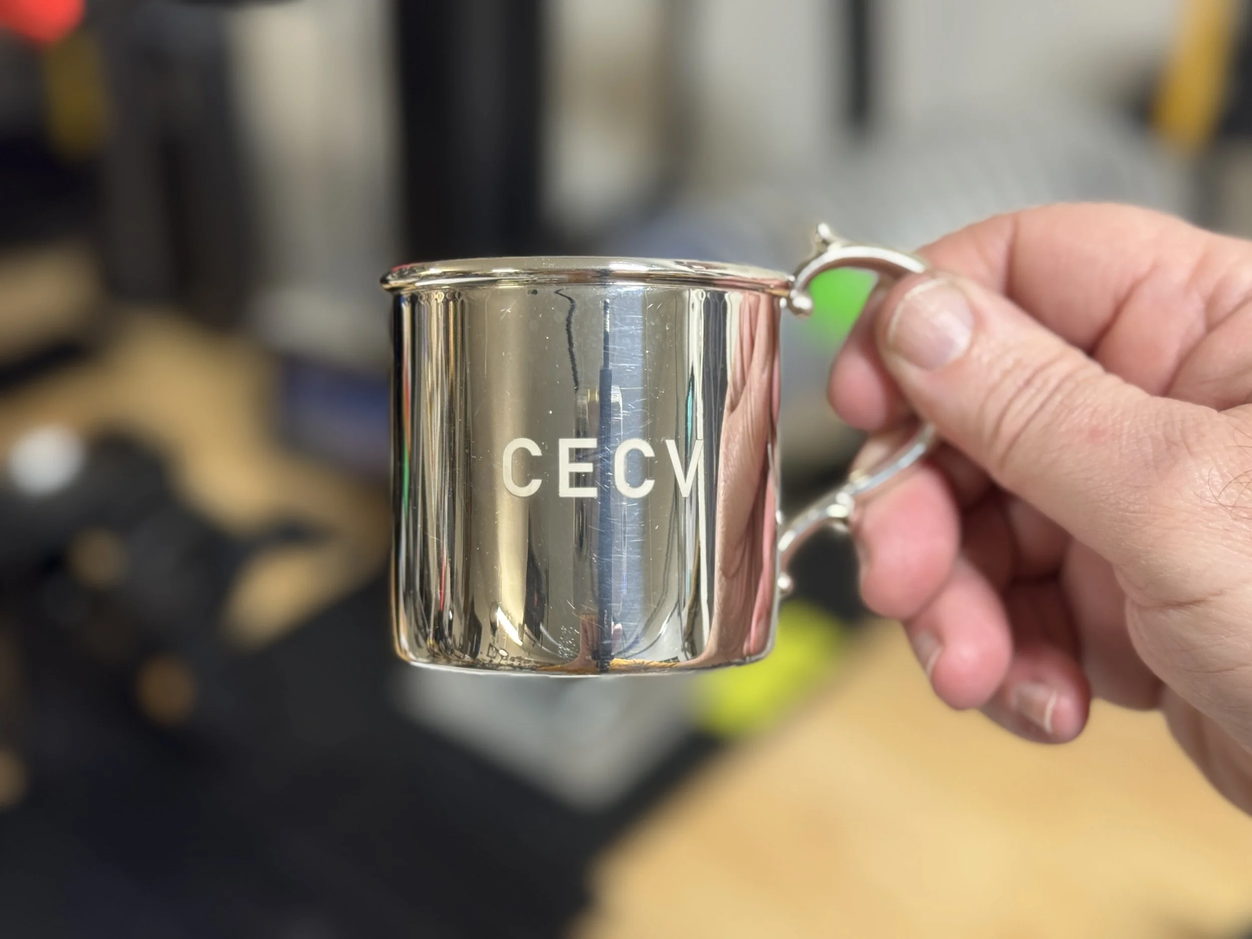 A hand holding a shiny silver mug with the letters 'CECM' on it, in an indoor setting with blurred background.