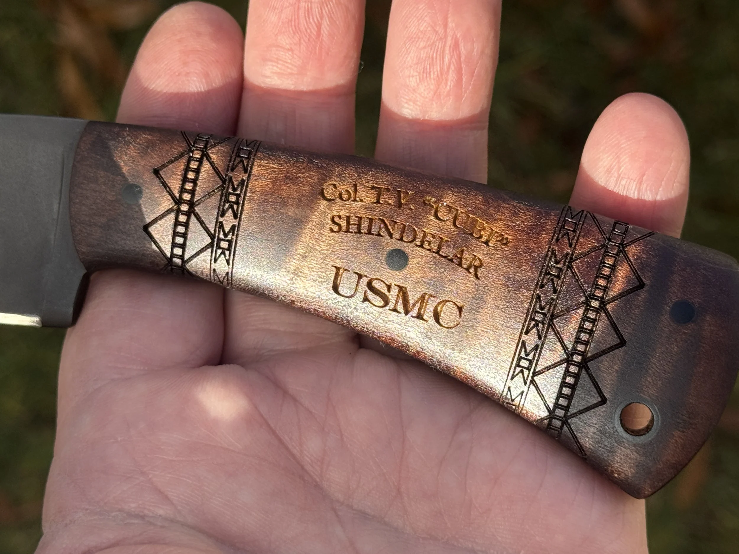 Close-up of a hand holding a military-style fixed blade knife with a metallic handle engraved with text and decorative patterns, showing a weathered finish.