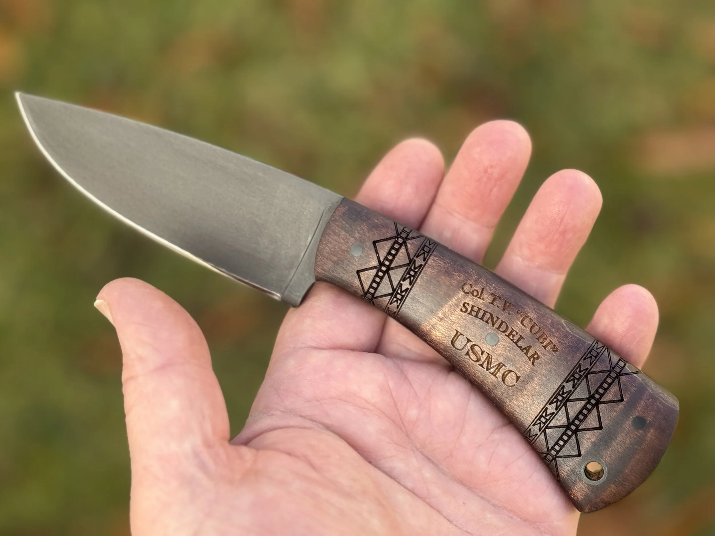 A hand holding a fixed-blade knife with a wooden handle engraved with geometric patterns and the words "COLT V. GURP SHINDELAAR USMC".