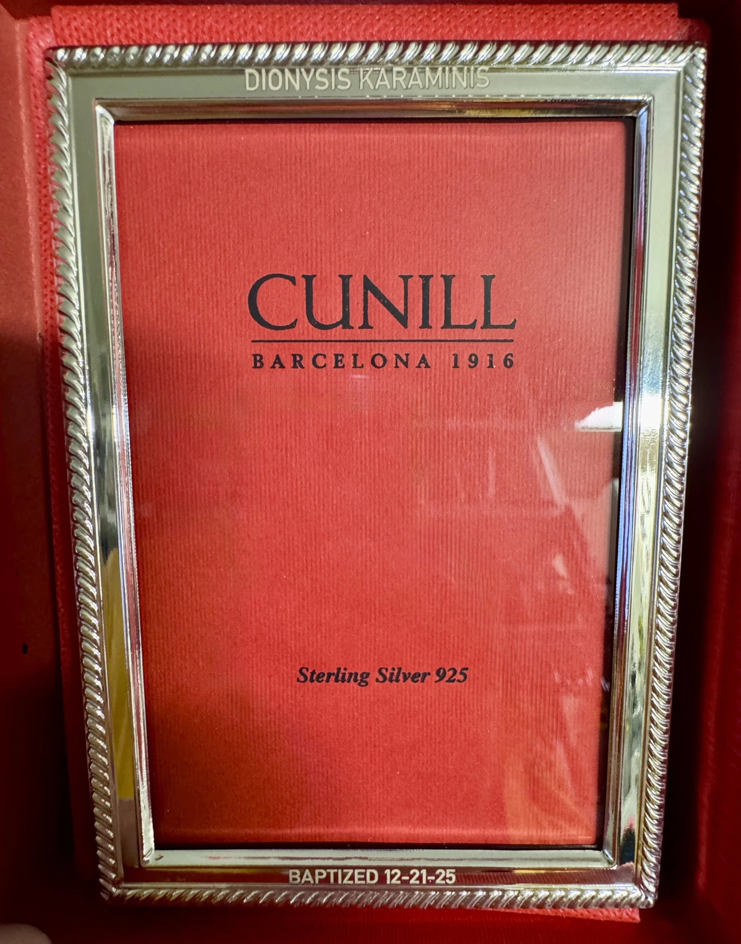 A silver picture frame with text inside that reads "CUNILL BARCELONA 1916 Sterline Silver 925"; at the top, the frame reads "DIONYSIS KARAMINIS" and at the bottom, it reads "BAPTIZED 12-21-25." The background inside the frame is red.