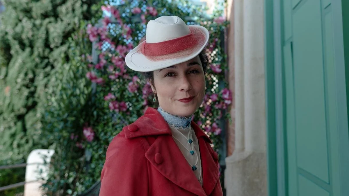 A woman wearing a white hat with a pink band, a red coat, and a lace collar, standing outdoors with pink flowers and green foliage in the background.
