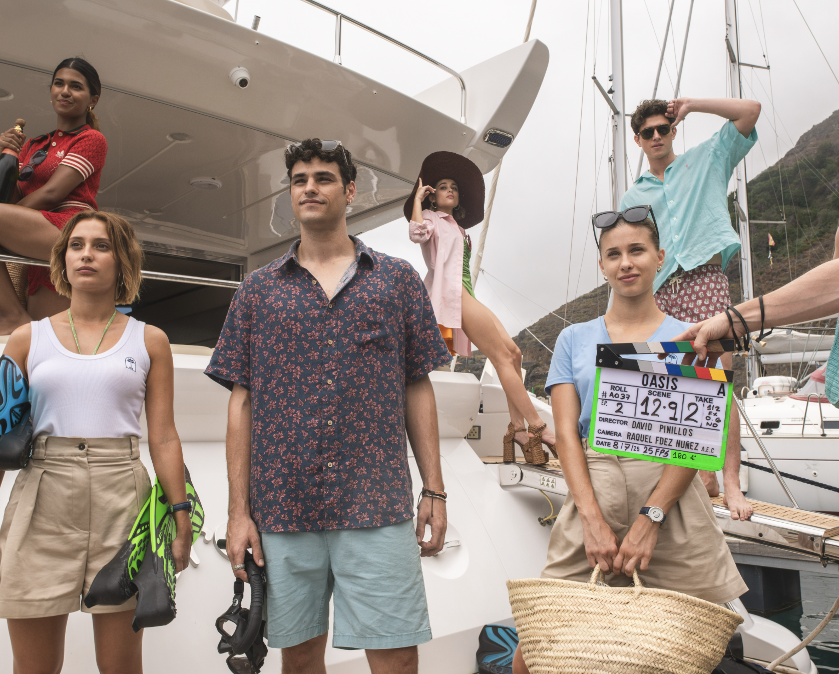 Group of young people standing on a yacht, holding scuba fins and a clapperboard labeled 'Oasis' with scene details, dressed in casual summer clothing, some with sunglasses, with sailboats and mountains in the background.