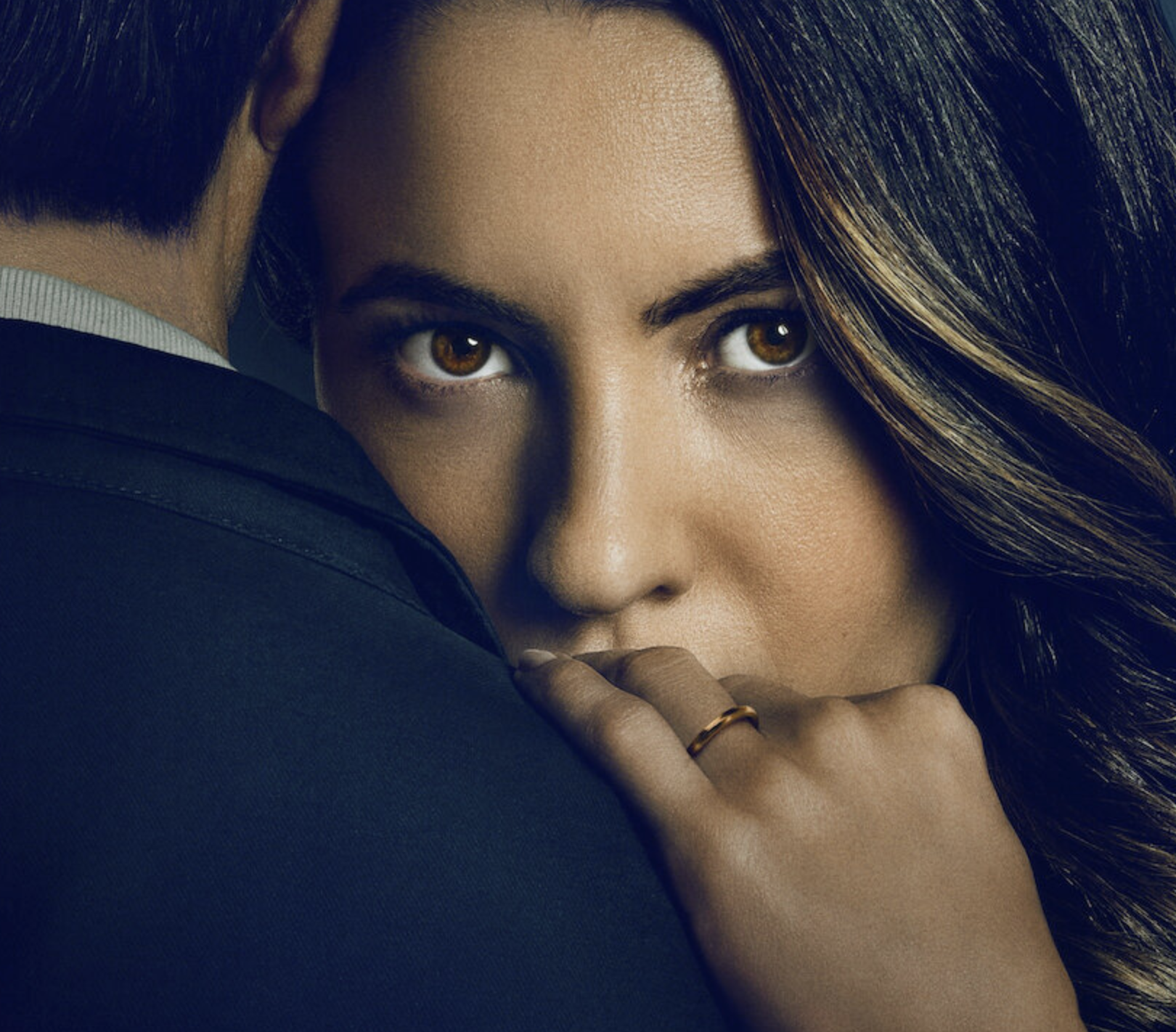 Close-up of a woman with brown eyes and wavy hair, partially hidden behind a man in a suit, with her finger near her lips.