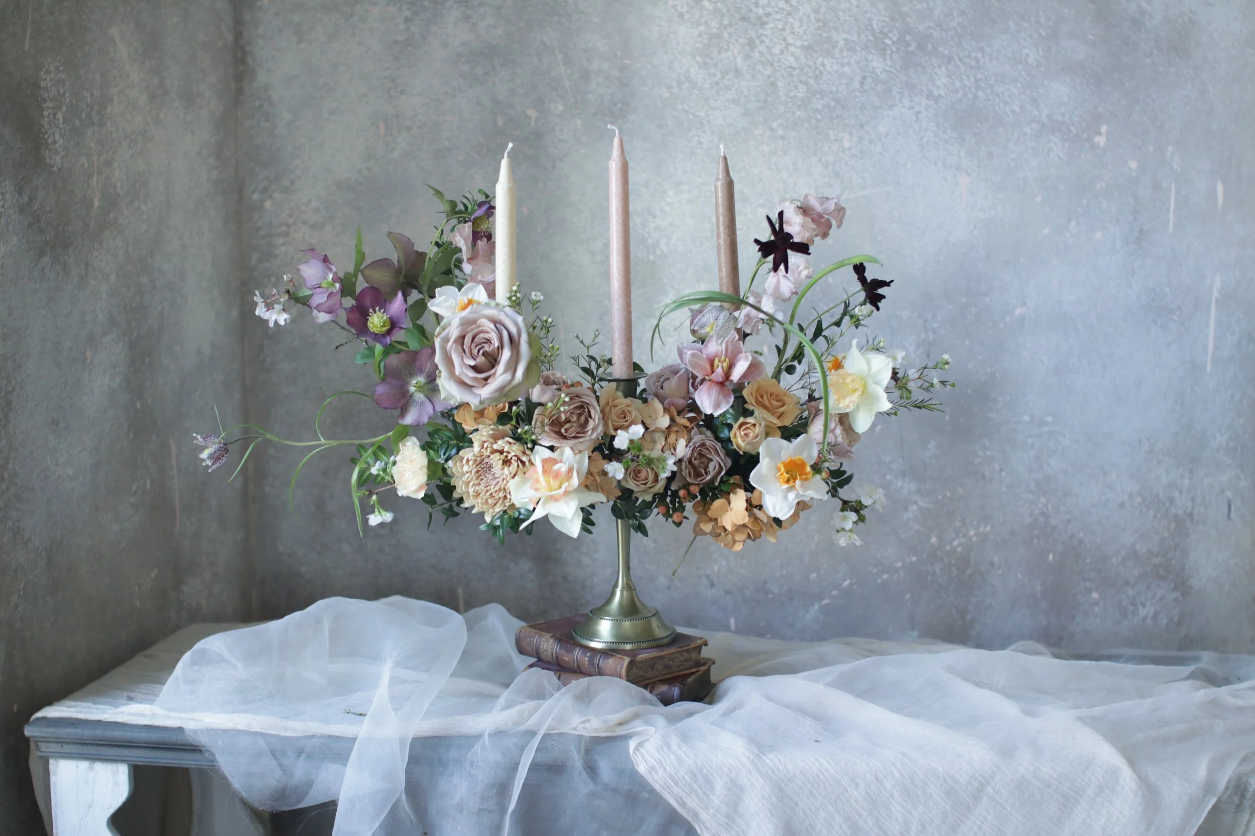 A floral centerpiece with pink, white, and purple flowers, including roses and orchids, in a brass vase with three tall candles on top, placed on a wooden book on a white draped table against a grey wall.