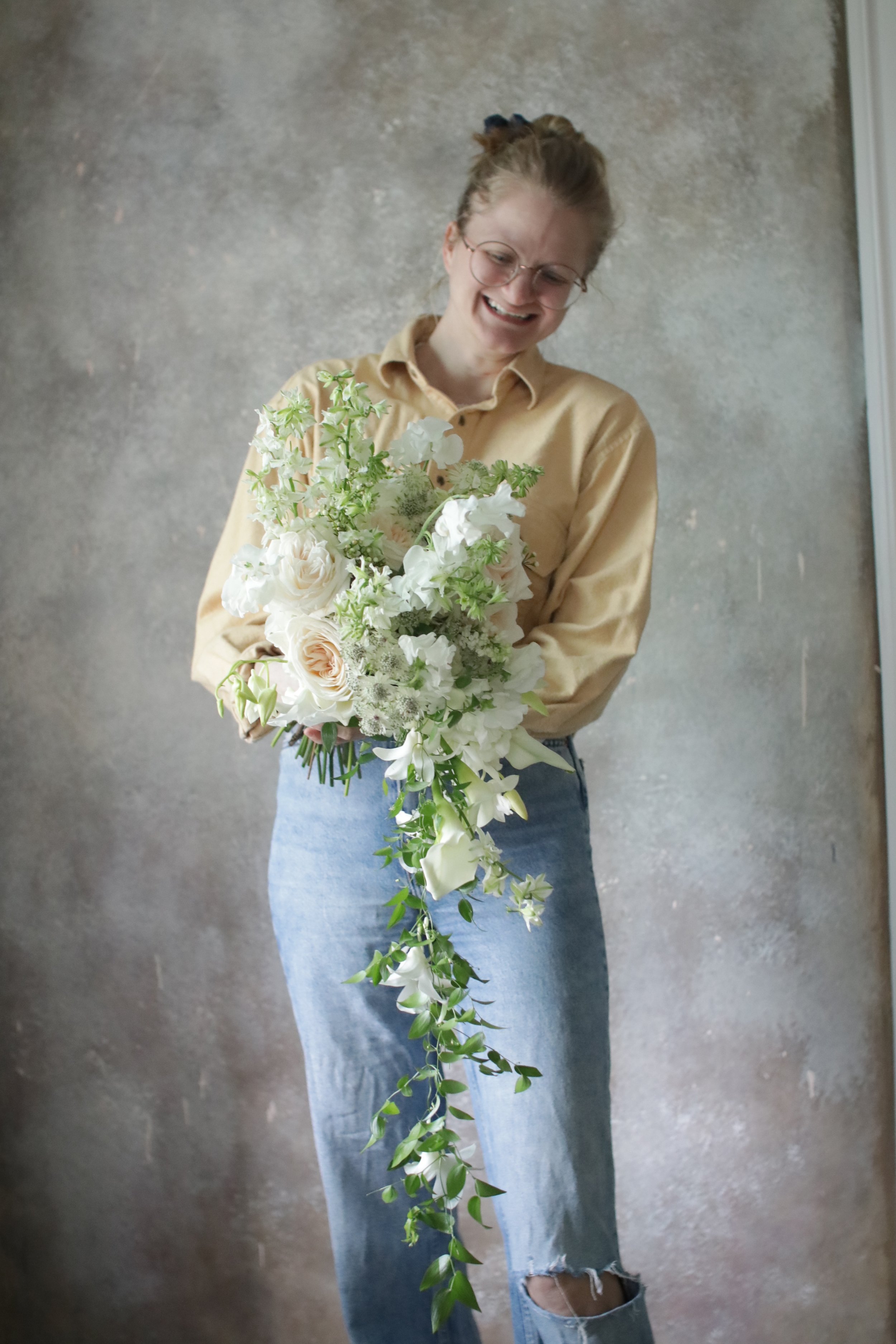 A woman smiling and holding a cascading bouquet of white and green flowers against a neutral textured background.