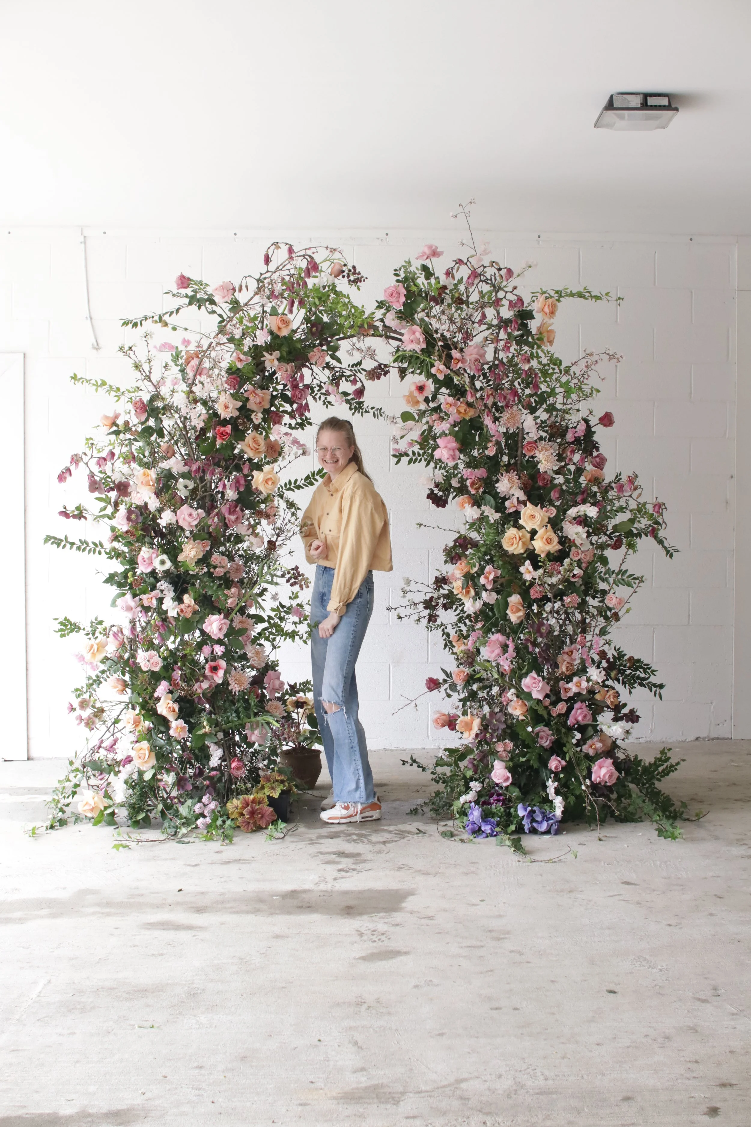 A woman smiling and standing in front of a large floral arch made of pink, peach, purple, and white flowers, set against a white brick wall in a minimalistic room.