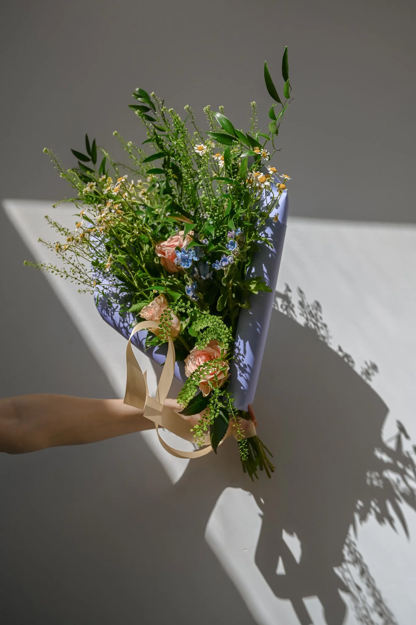 Hand holding a bouquet of mixed flowers with greenery in a lavender wrapping, casting shadows on a white surface.