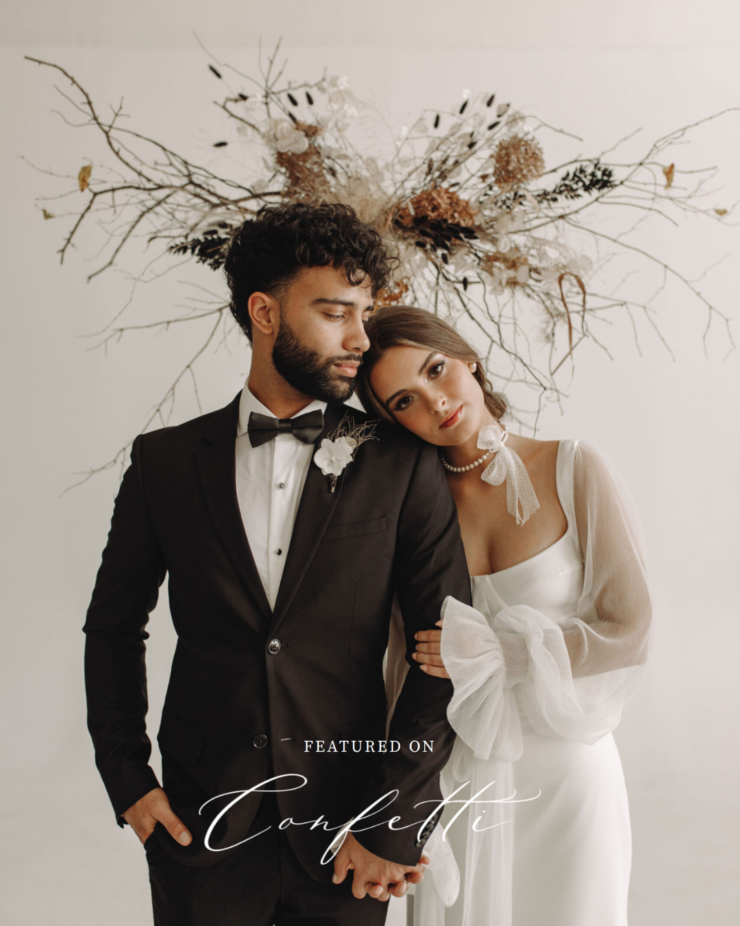 A bride and groom in wedding attire, standing close together against a plain wall with a decorative dried floral arrangement behind them, holding hands and looking at each other softly.