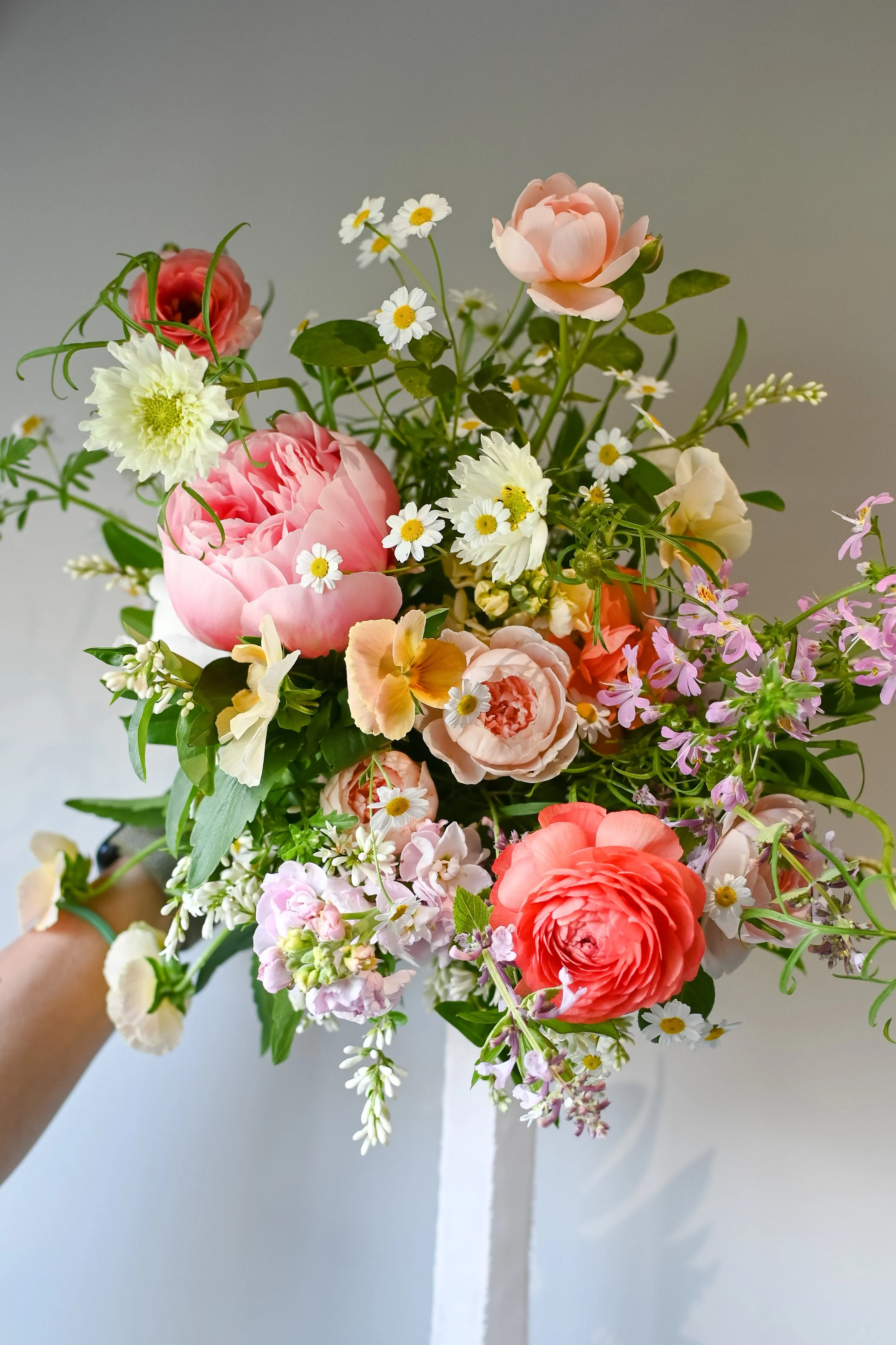 A colorful bouquet of various flowers including pink peonies, white daisies, and other mixed flowers, held by a person's hand against a neutral background.