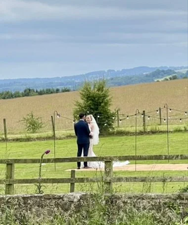 A bride and groom standing in a field in front of a fence, with a countryside landscape in the background, featuring hills and a cloudy sky.