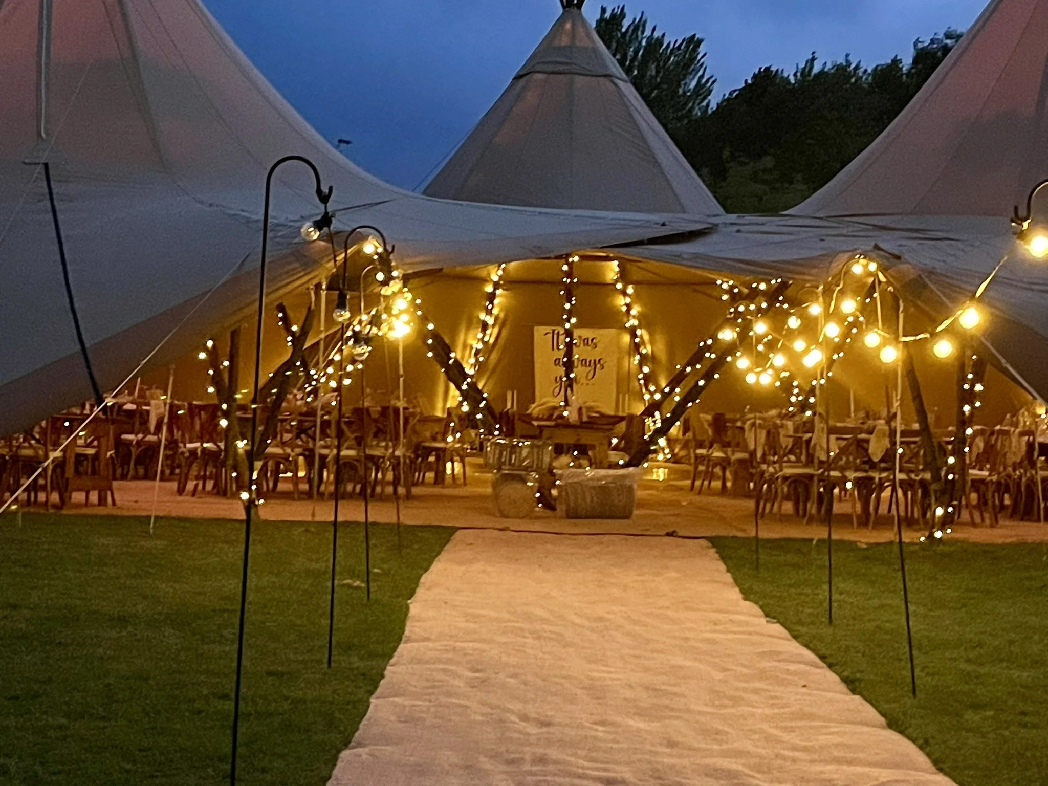 Outdoor tented event with string lights, tables, and chairs on grass.