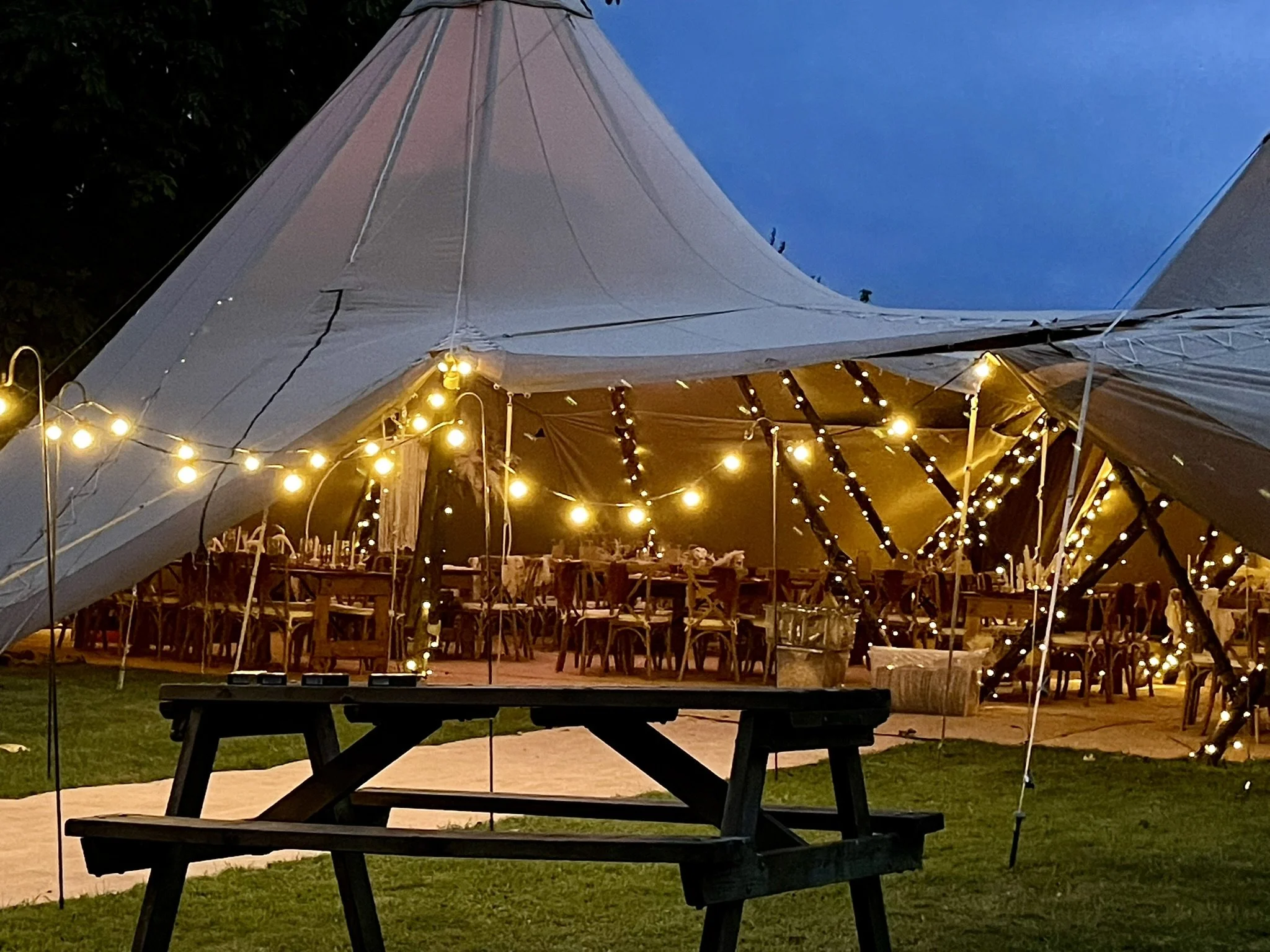 Outdoor event tent with string lights and wooden tables and chairs set up for a gathering, with a picnic table in the foreground.