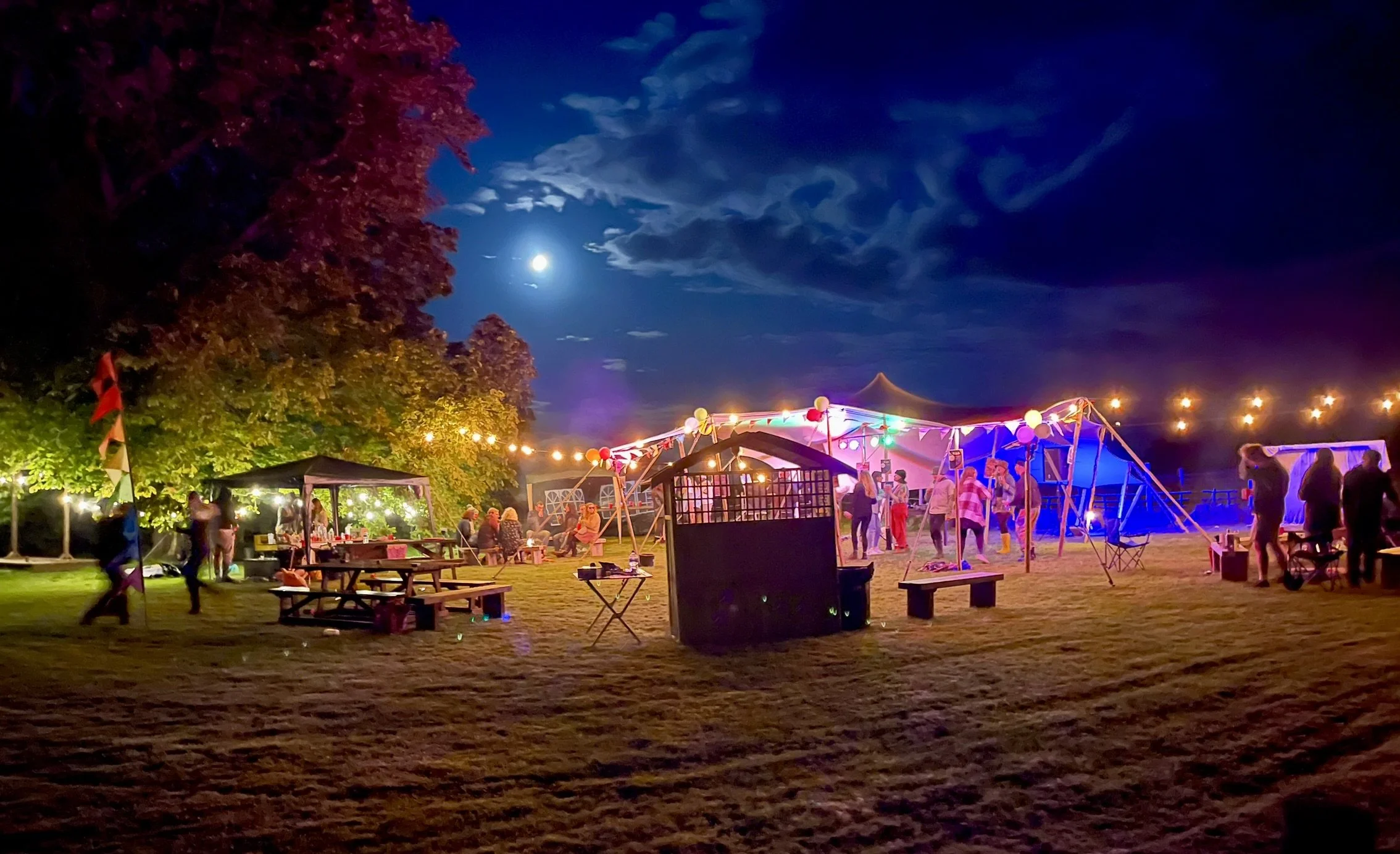 Nighttime outdoor party with colorful lights, tents, and people gathering on a lawn with a full moon visible in the sky.