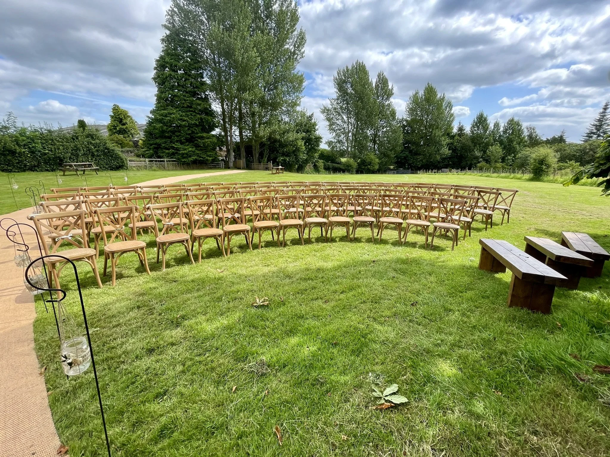 Outdoor wedding ceremony setup with wooden chairs and benches on a grassy lawn under a partly cloudy sky.