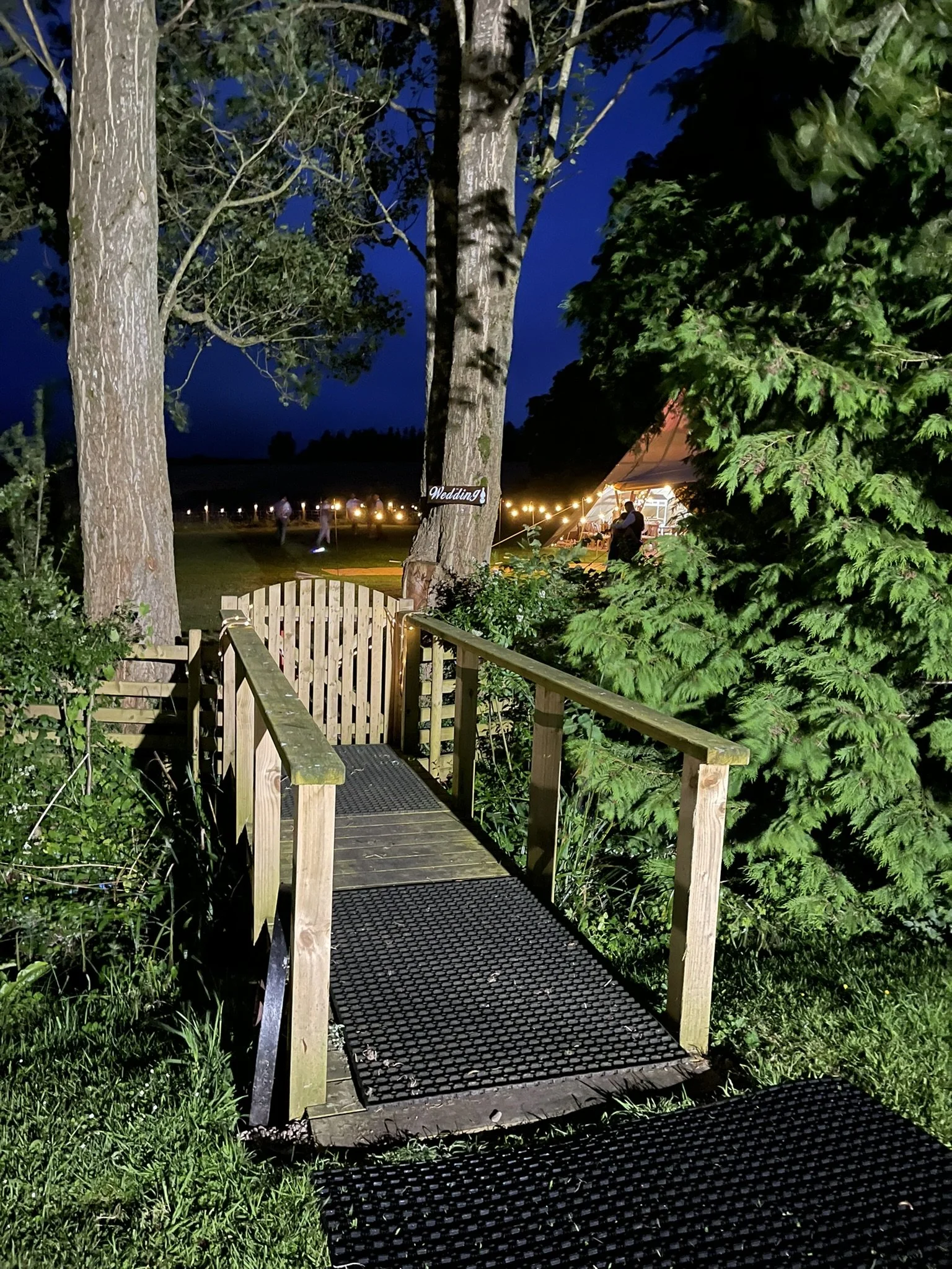 Small wooden bridge with railing leading to a garden, surrounded by trees and foliage, with a wedding sign and tent with lights visible in the background at night.