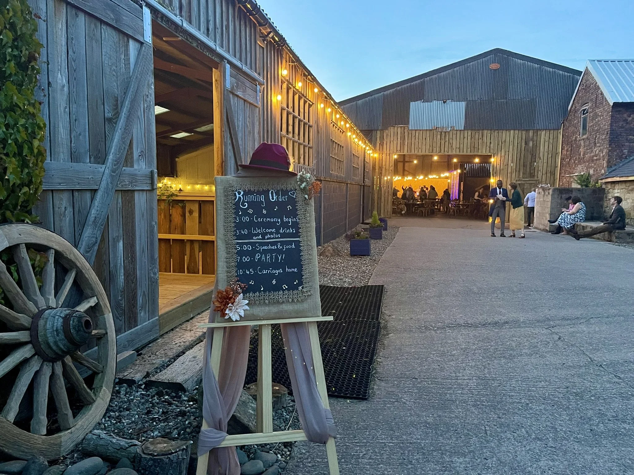 Rustic barn wedding venue with open doors revealing interior lights. A chalkboard sign displays the event schedule next to a wooden wagon wheel. Guests gather outside the barn, which is adorned with string lights. Evening setting.