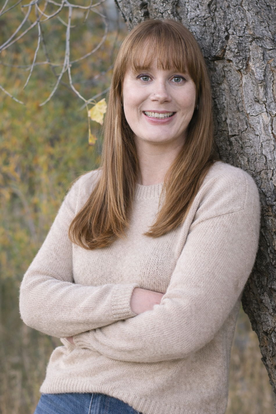 A woman with long auburn hair and bangs, smiling and standing outdoors leaning against a tree with crossed arms, wearing a cream-colored sweater during autumn.