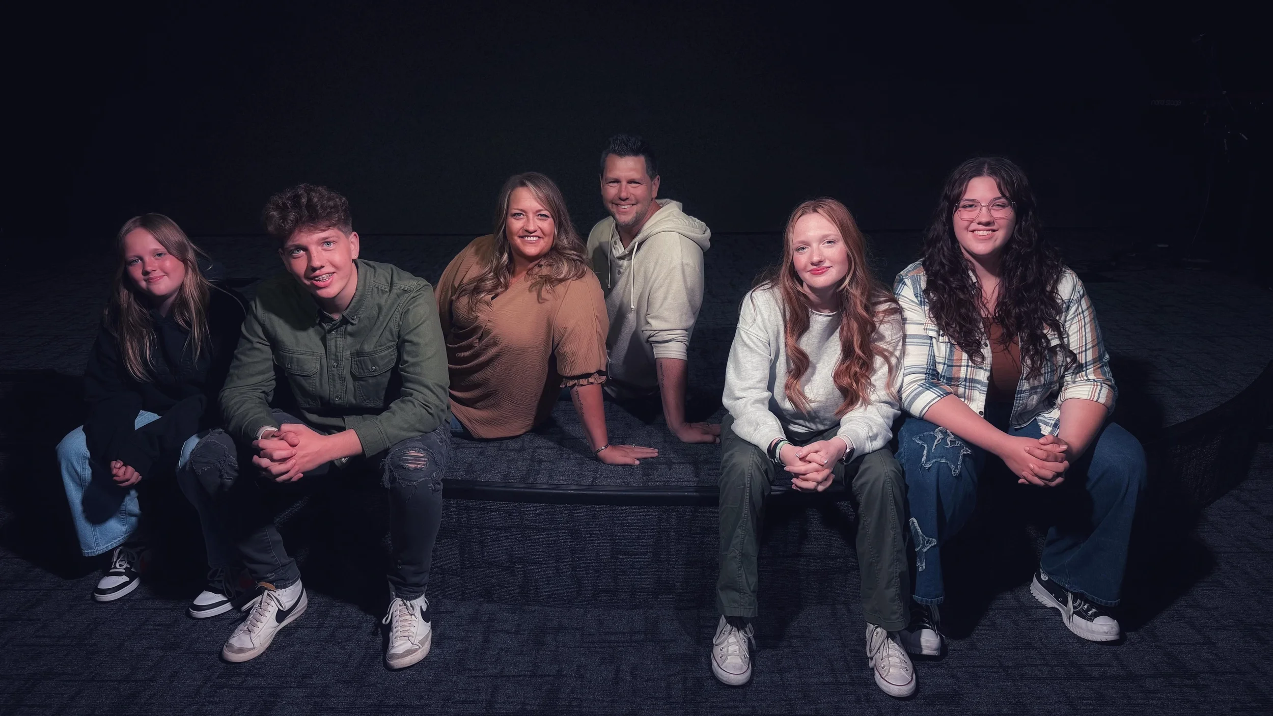 Group of six people sitting on a dark stage or platform, smiling at the camera.