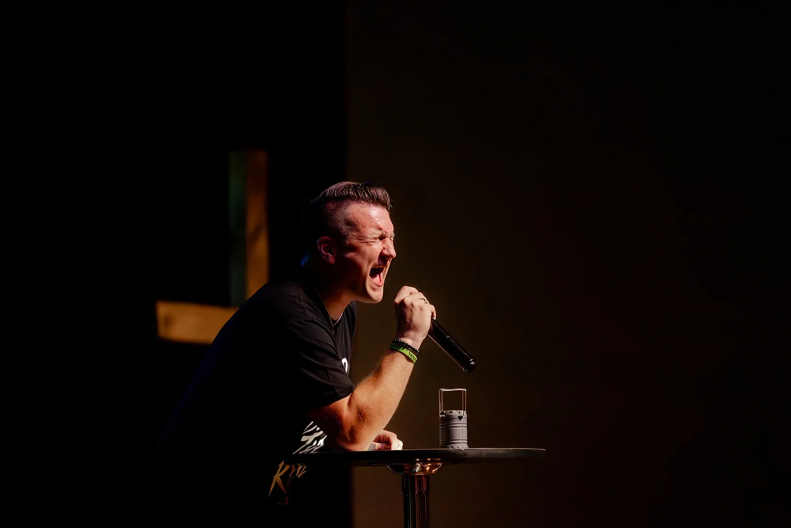 A man with short dark hair passionately singing into a microphone on stage, illuminated by a spotlight, with a dark background.
