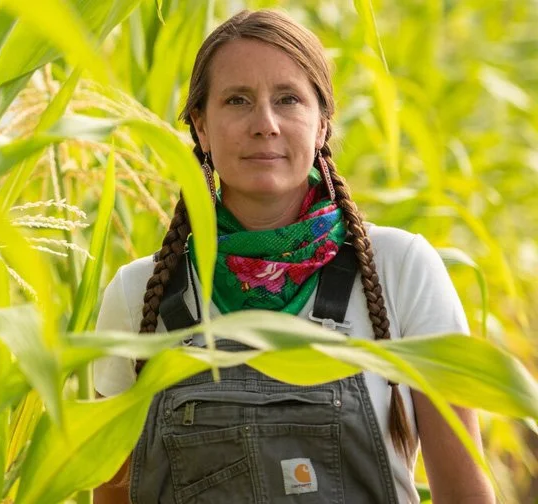 A woman with braided hair and a green, pink, and purple scarf around her neck standing among tall green plants.