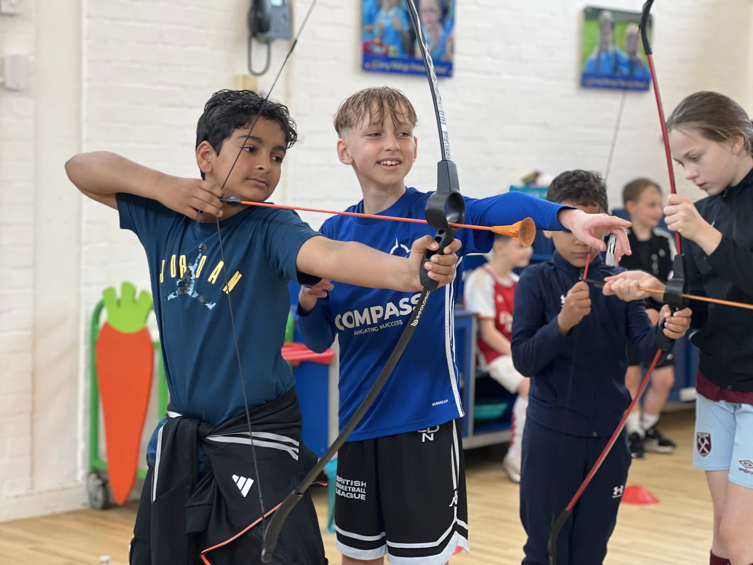 Children practicing archery indoors, aiming with bows and arrows. They appear focused and involved in the activity with bows drawn. Other children are observing or preparing in the background.