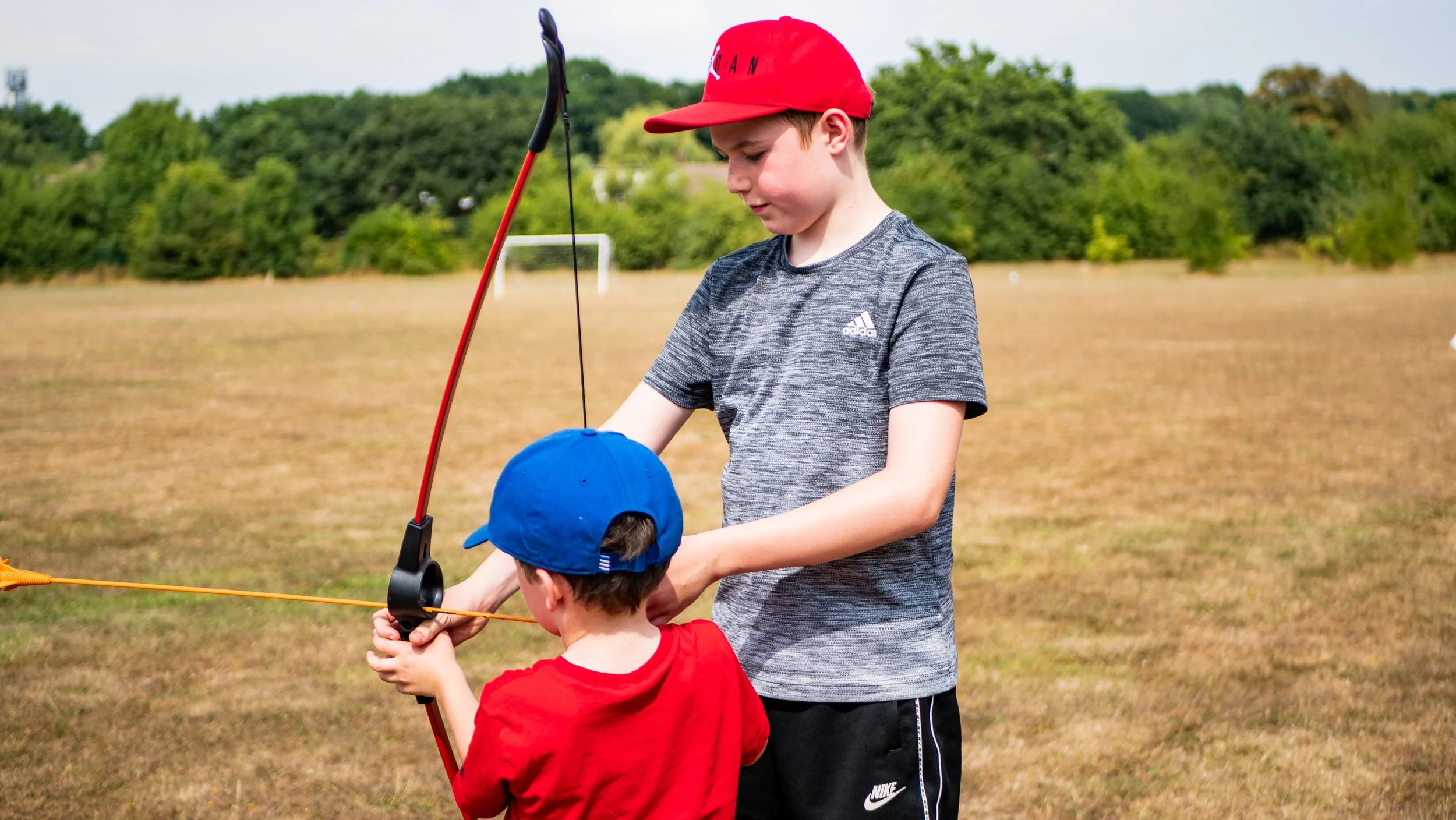 Two boys practicing archery outdoors. One boy is assisting the other with aiming a bow and arrow. They are standing on a dry grassy field, with trees in the background. Both are wearing casual clothing and caps, while the younger boy is holding the bow.