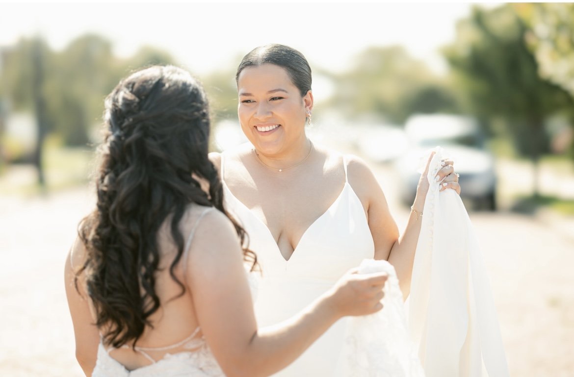 Two women in wedding dresses smiling outdoors, one with dark hair holding a white garment, with trees and cars in the background.