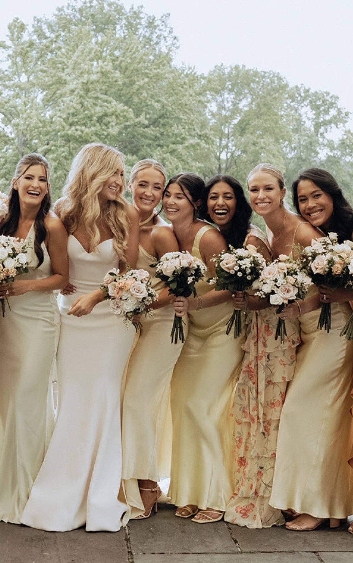 Group of women in cream-colored dresses holding bouquets of flowers, posing outside in front of trees, smiling for a wedding photo.