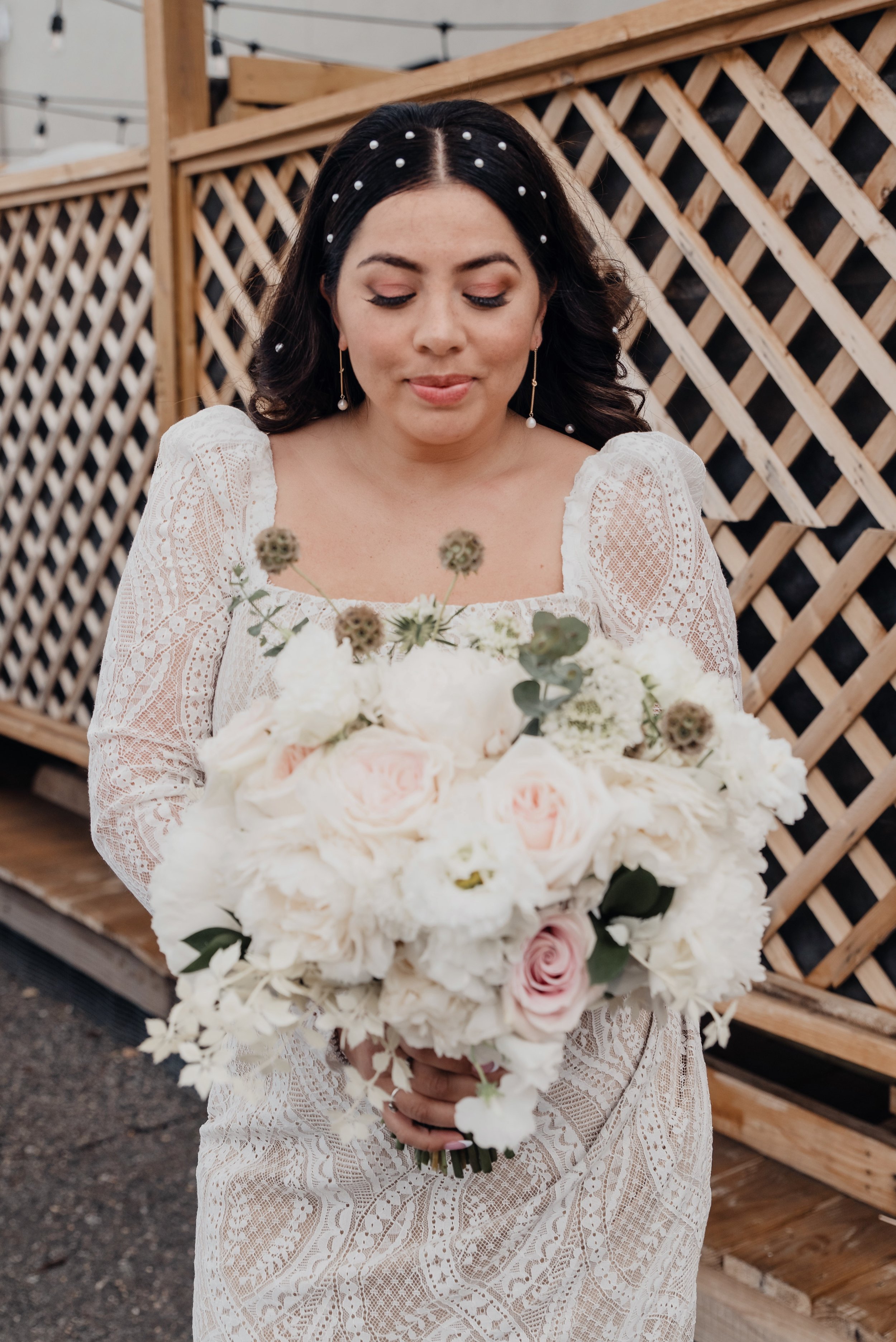 A bride holding a bouquet of white and pink roses, with a wooden lattice fence in the background.