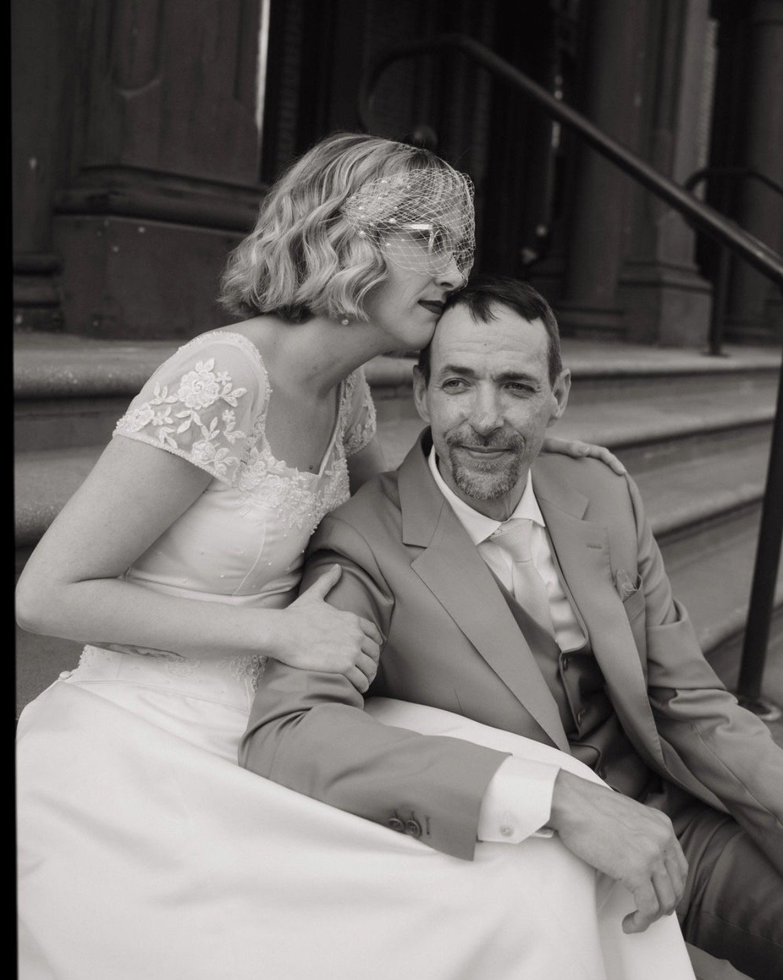 A bride and groom sitting on steps outside a building. The bride is wearing a wedding dress with floral embroidery and a birdcage veil. The groom is in a suit with a tie. The bride is gently kissing the groom's head, and they appear happy.