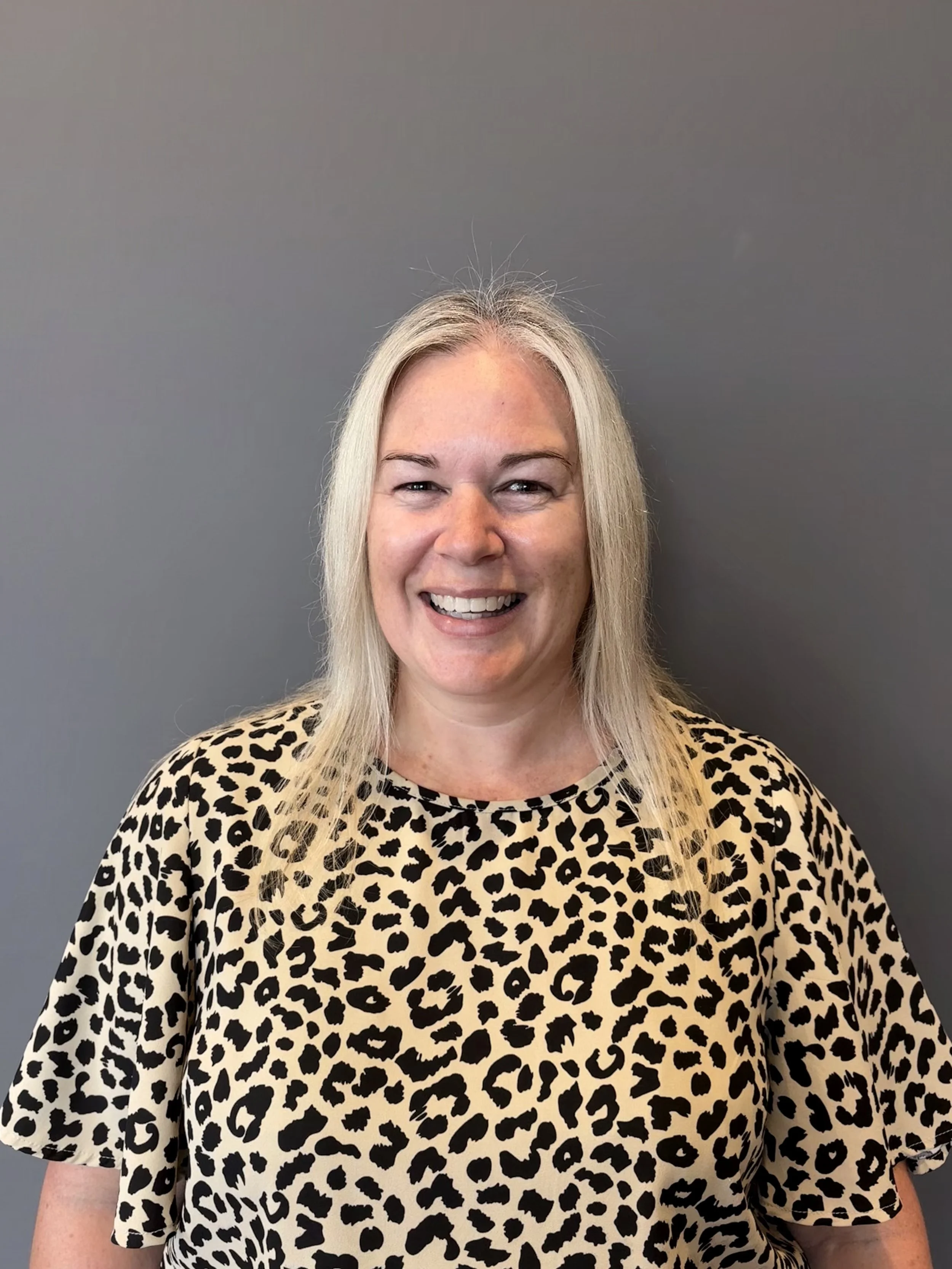 A smiling woman with shoulder-length blonde hair wearing a leopard print top against a plain white background.