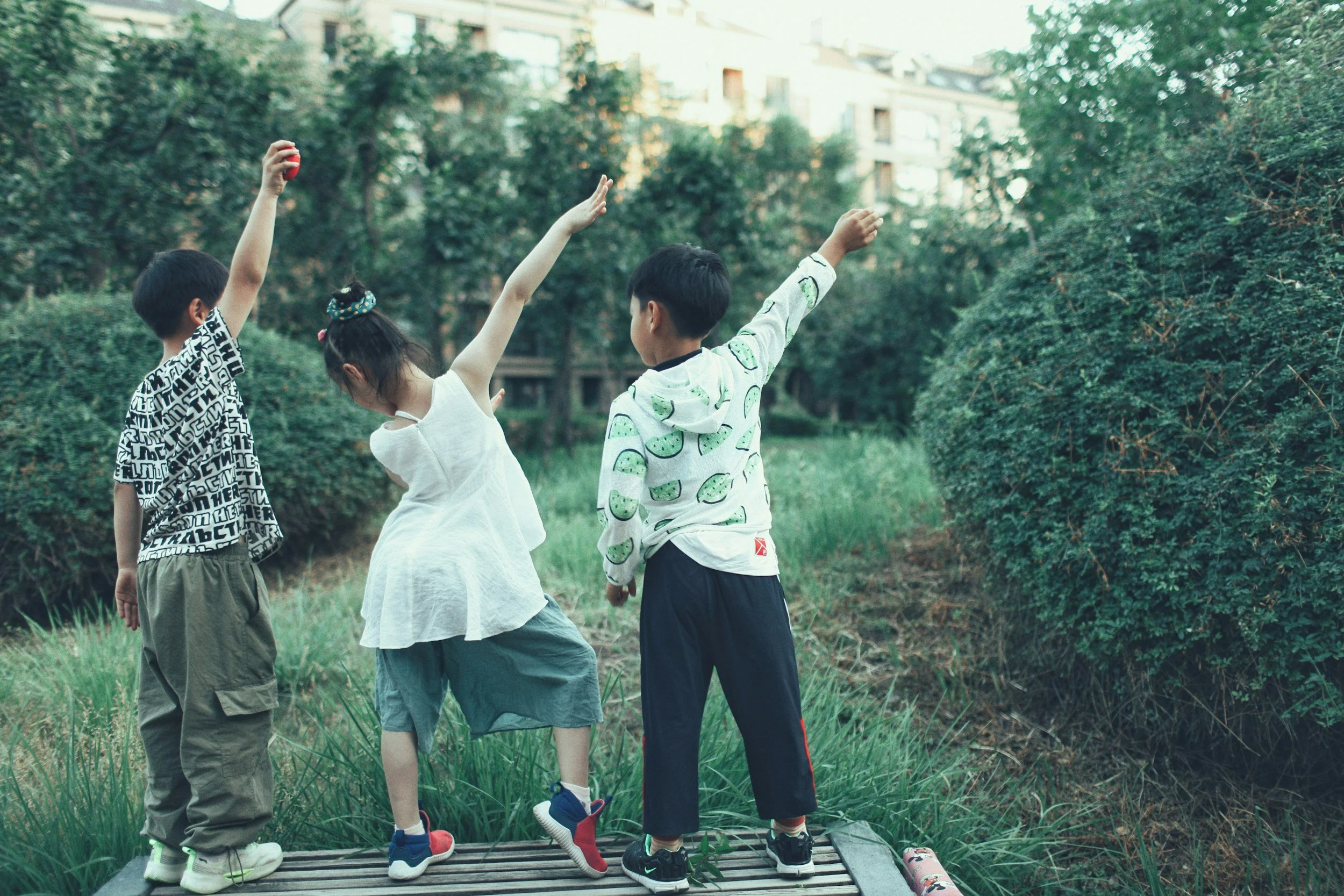 Three children standing on a wooden platform in a park, facing away, raising one arm. They are surrounded by greenery with trees and bushes, and buildings are visible in the background.