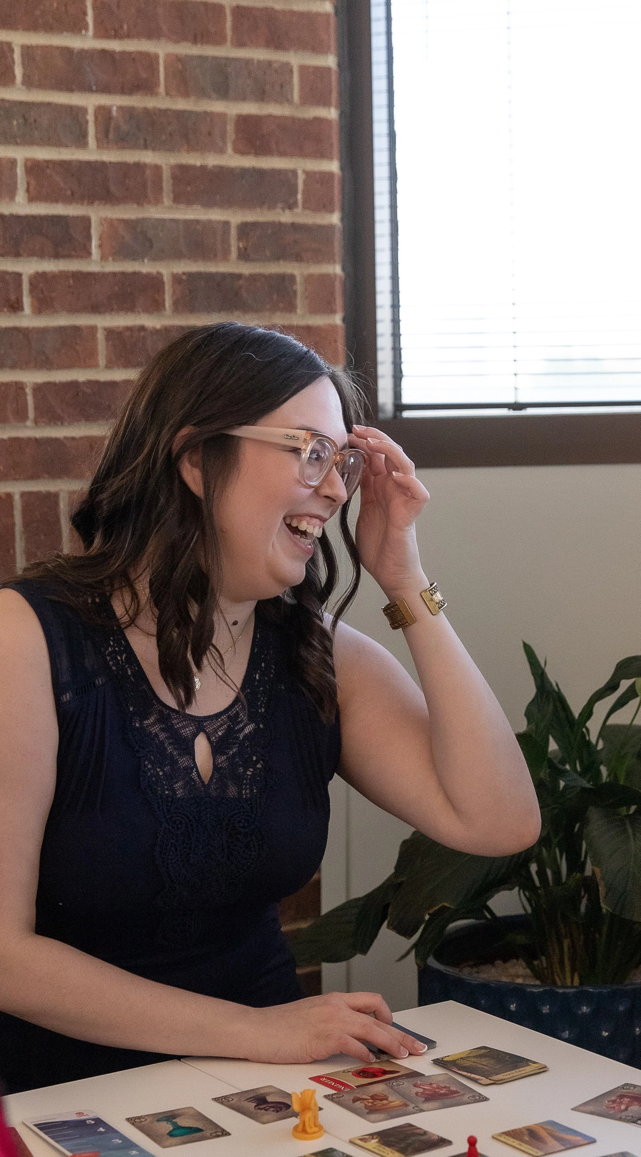 A young woman with dark hair and glasses playing a tabletop card game, smiling and touching her forehead, with cards and game pieces in front of her in an indoor setting with a brick wall and window blinds.