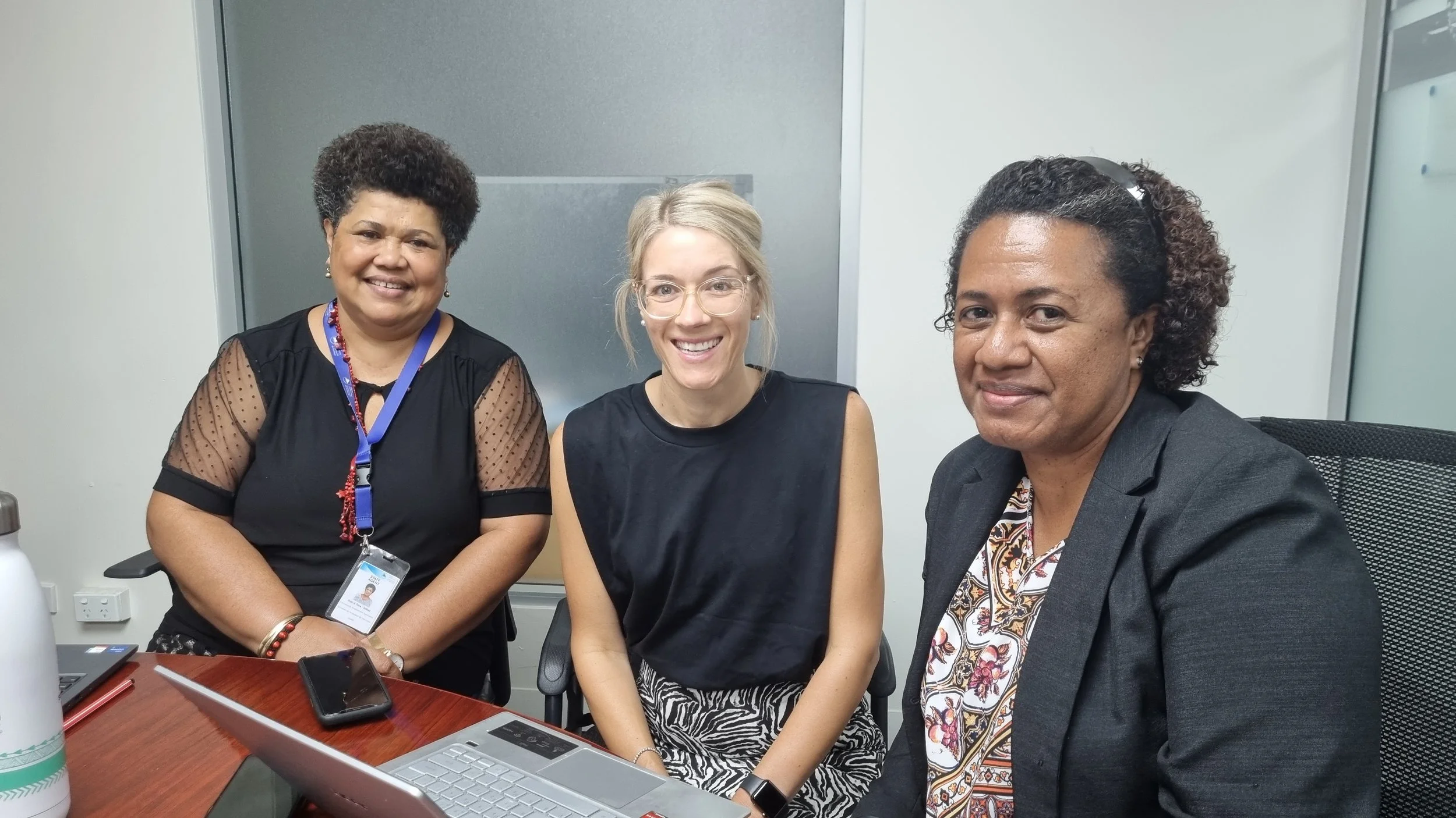 Three women sitting at a table in an office setting, smiling at the camera. Laptops and a water bottle are on the table.