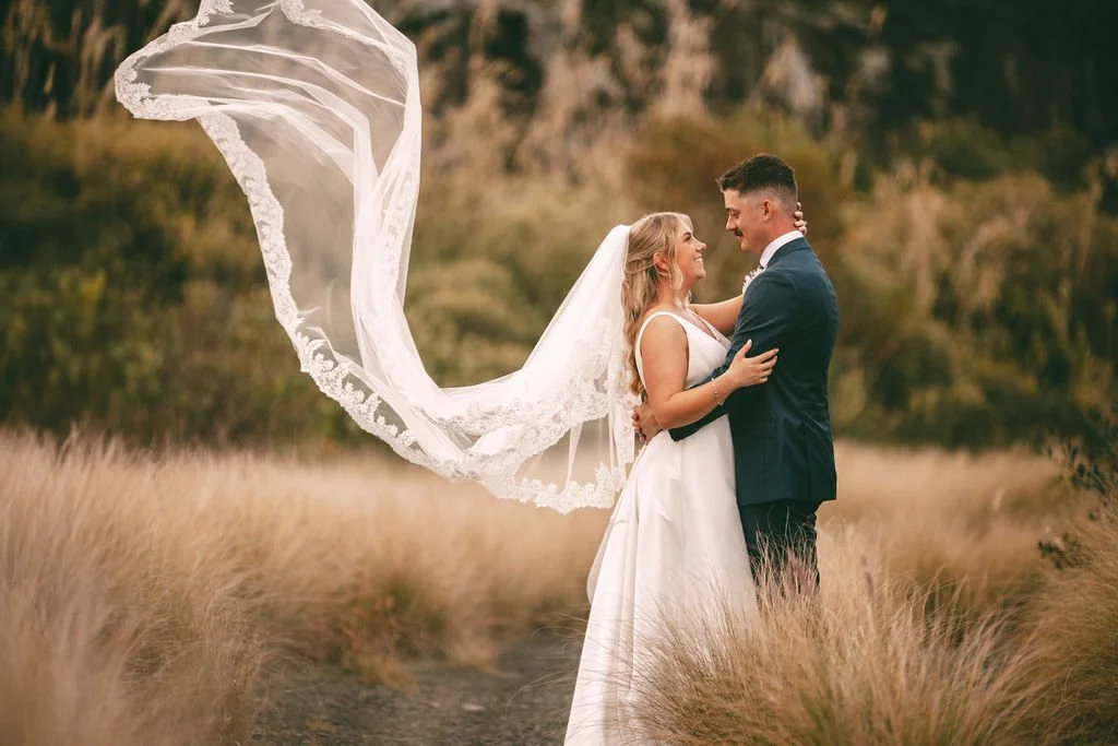 Charlotte and Ethan embracing in a field with her wedding veil blowing in the wind