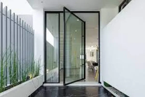 Modern glass pivot door leading into an indoor space, with a small balcony or patio area outside, featuring a vertical metal fence and potted plants.