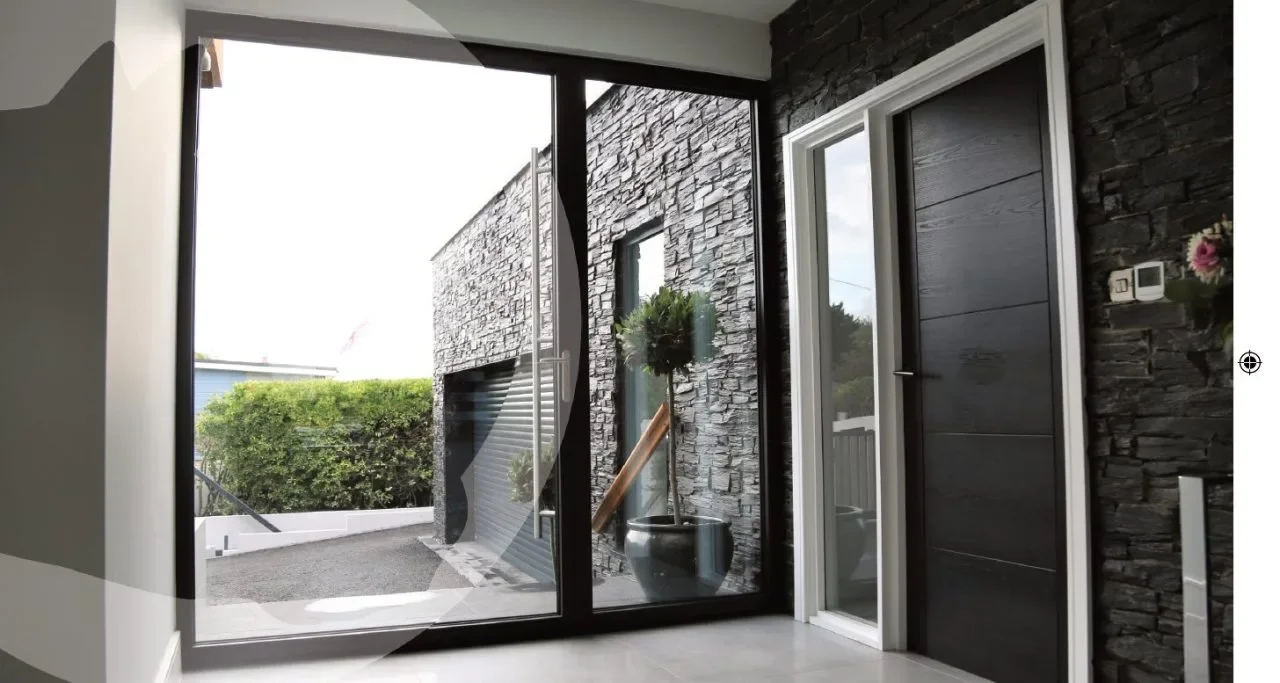 Interior view of a modern house with aluminium pivot glass door opening to a patio with bushes and a garage, featuring dark stone walls and a potted plant outside.