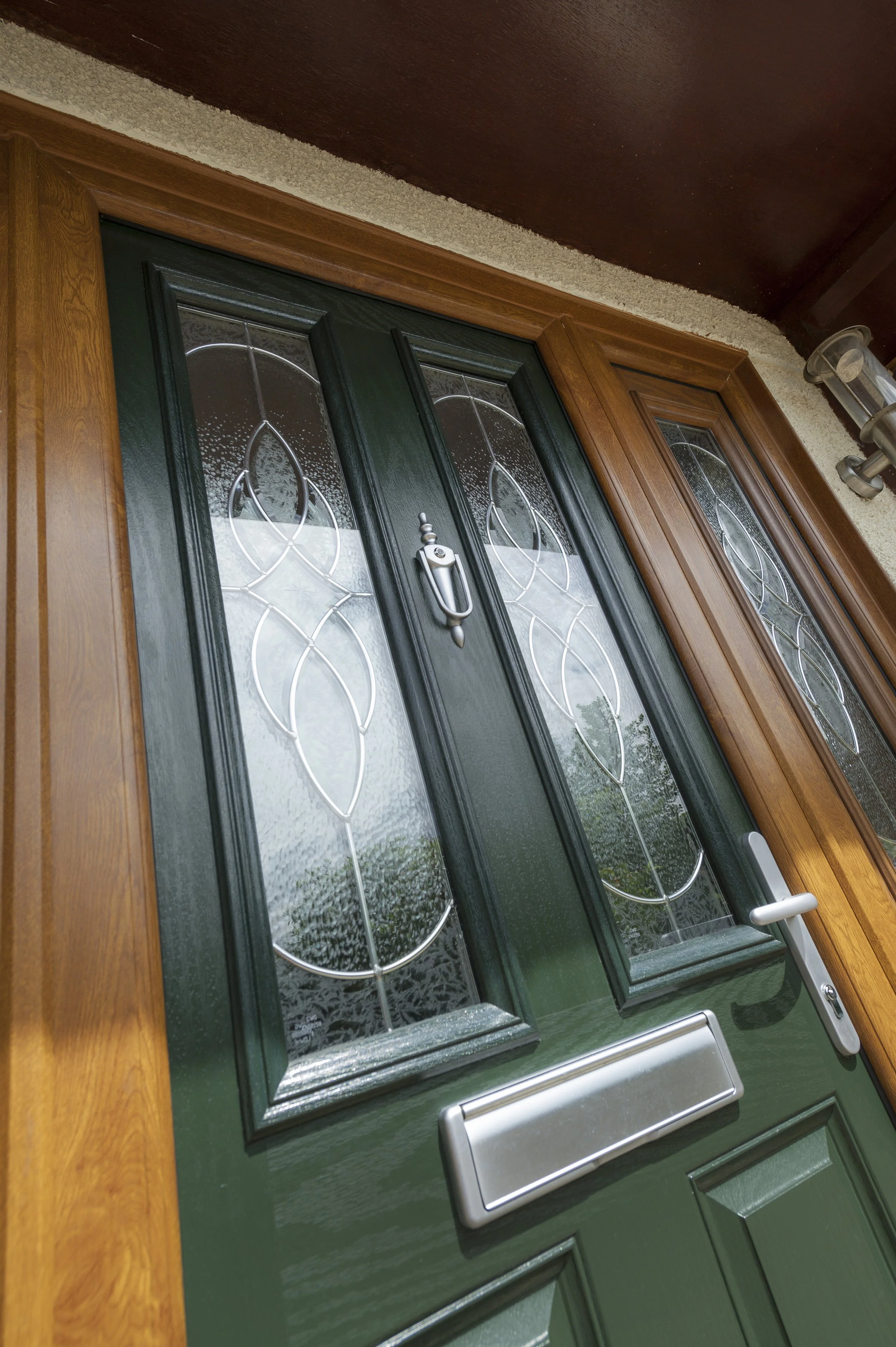 Front door with decorative glass panels, painted dark green, set in a wooden door frame, with a mail slot and handle, beside a wall-mounted light fixture.