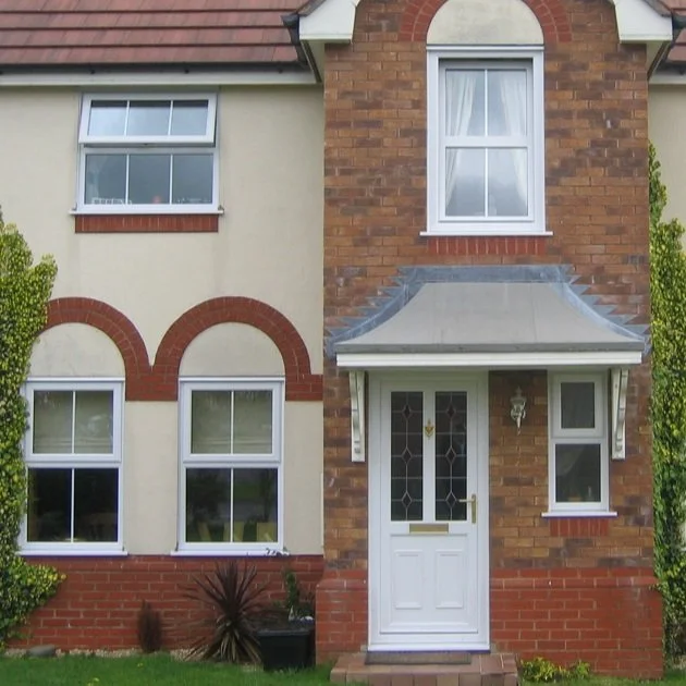 Front view of a two-story house with brick and beige exterior walls, white-framed windows, and a white front door with decorative glass, surrounded by green shrubbery.